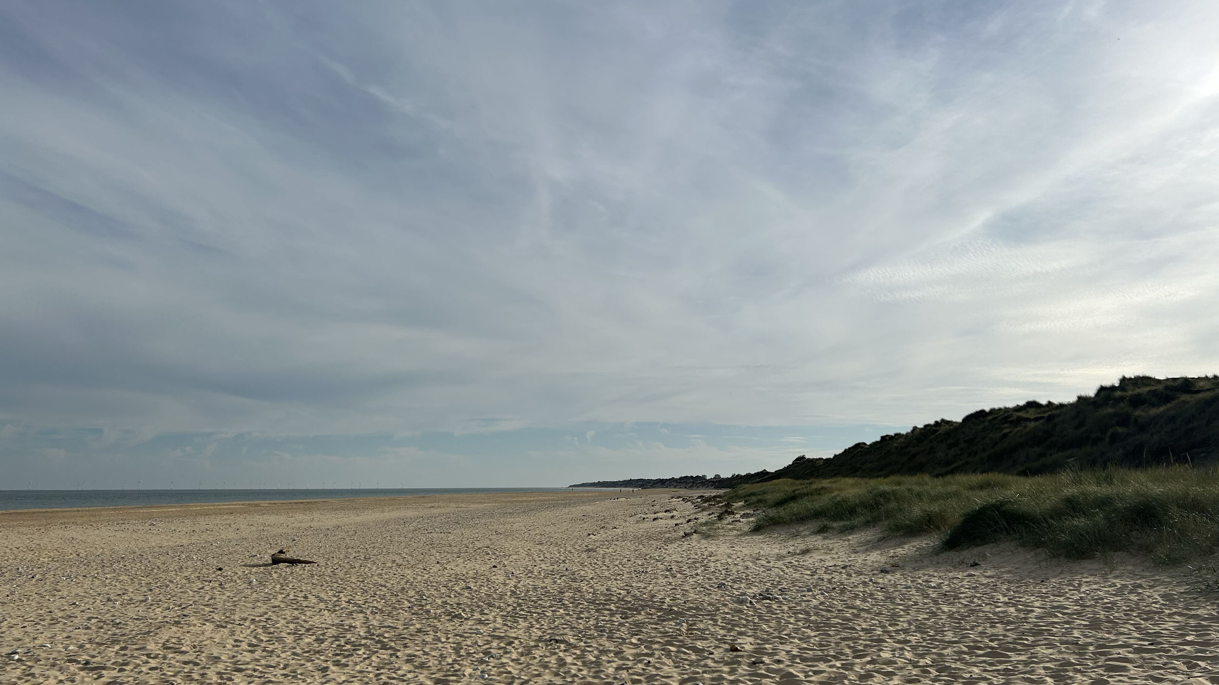 Empty sandy beach with grass-covered sand dunes on the right and a cloudy sky overhead.