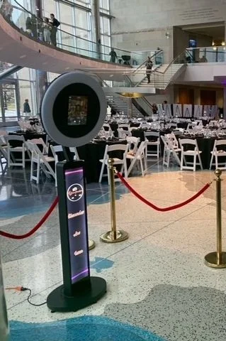 A large, circular photo booth with a touchscreen inside, set up in a spacious, modern event space with tables and chairs, a second-floor balcony, and a red velvet rope barrier.