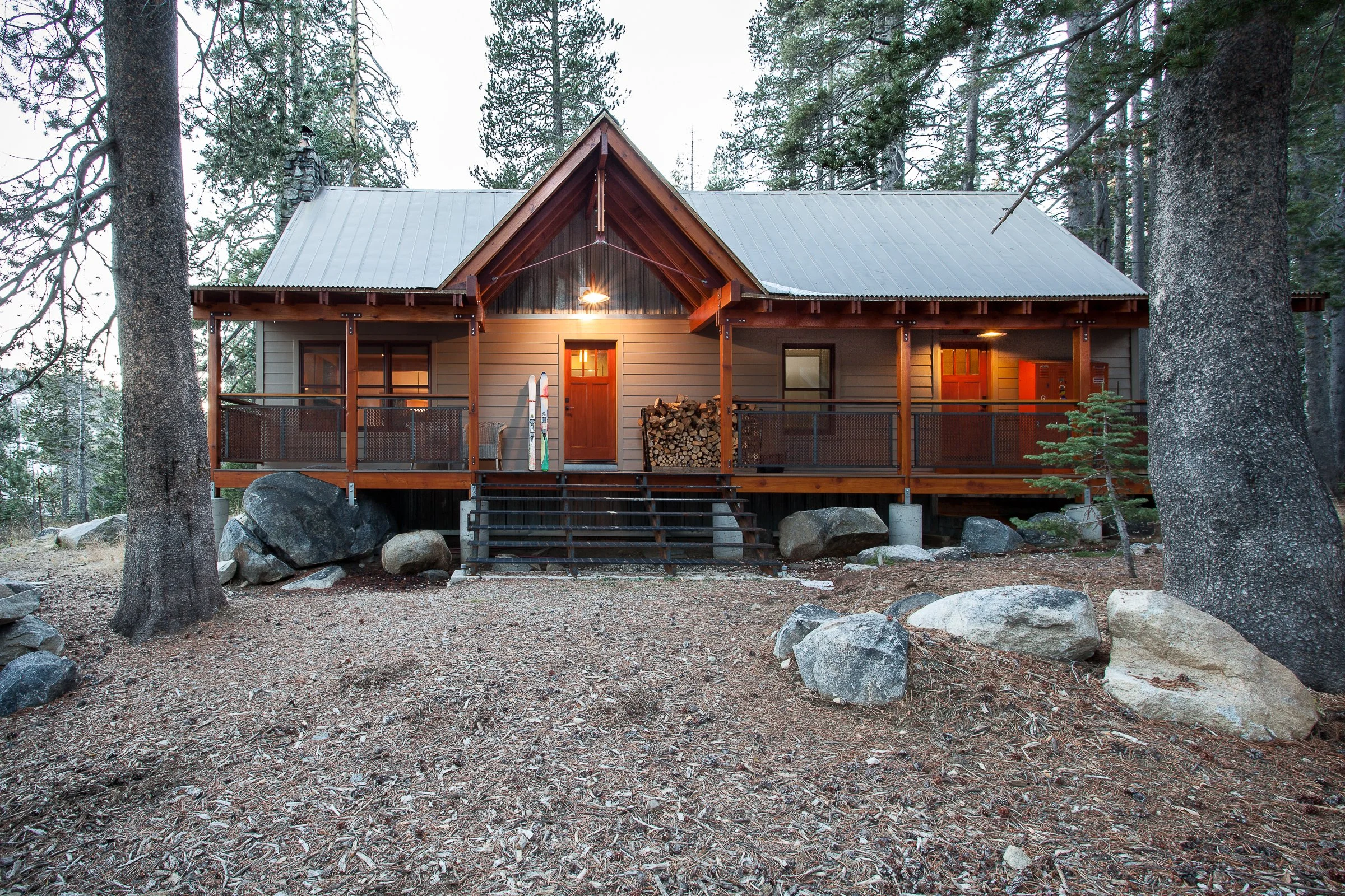 Rustic cabin surrounded by tall pine trees and rocky terrain, featuring a wooden porch and stacked firewood.