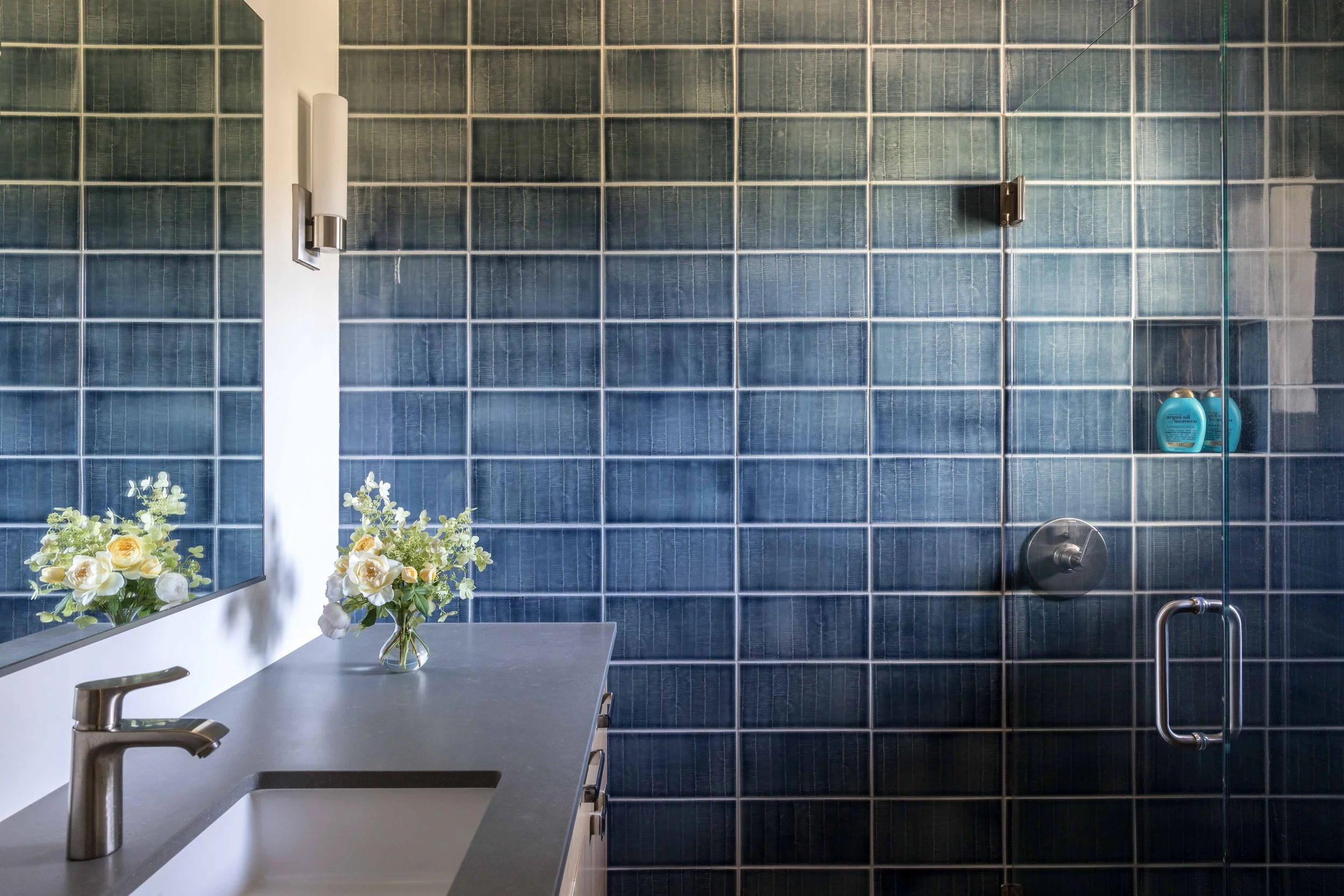 Modern bathroom with blue tiled walls, glass shower door, gray countertop, stainless steel faucet, and vase with flowers.