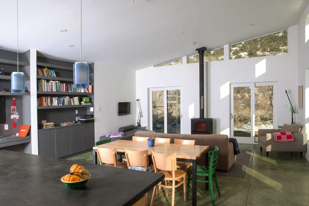 Modern living area with open kitchen, dining table, and wood-burning stove. Shelves with books and kitchen island with fruit bowl. Large windows and sliding doors with outdoor view.