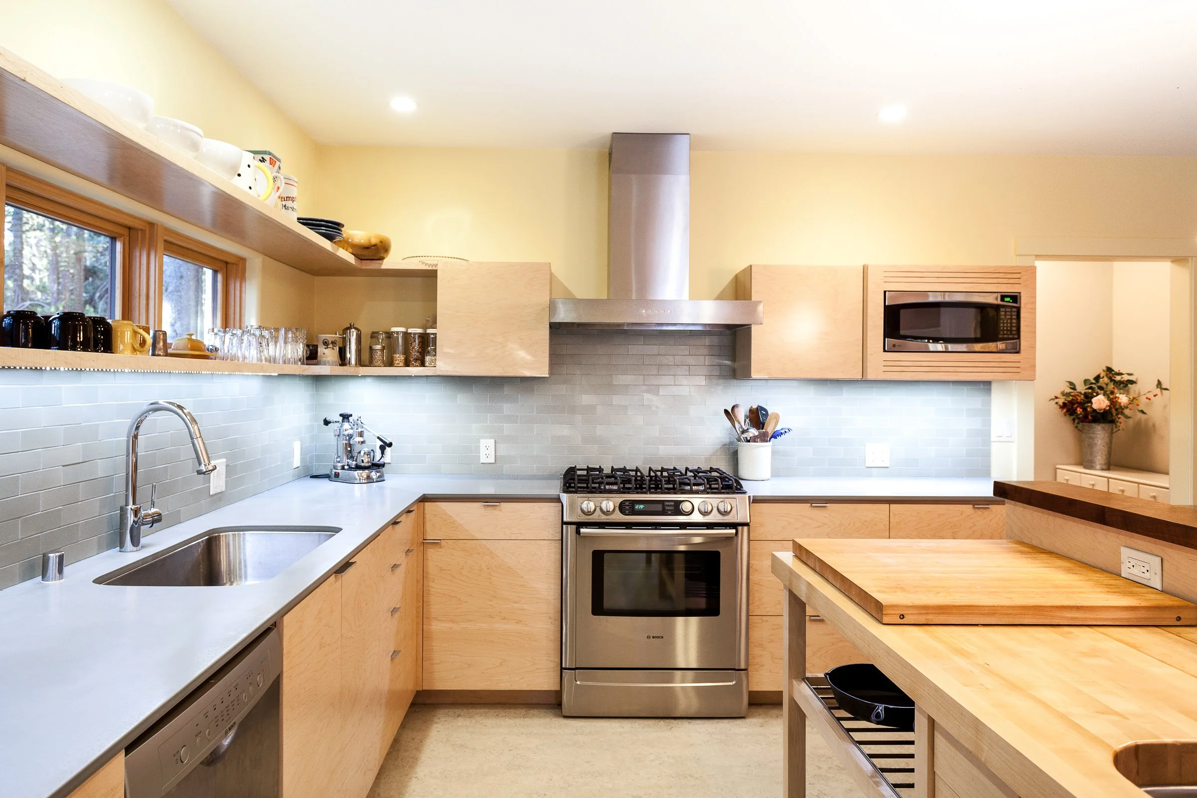 Modern kitchen with light wood cabinets, stainless steel appliances, a gas stove with a range hood, a built-in microwave, and a large kitchen island. The kitchen features a light tile backsplash, a stainless steel sink, and open shelving with dishes and glassware.