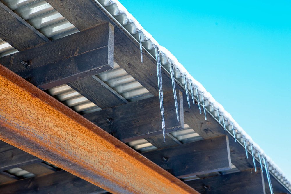 Close-up of icicles hanging from a metal roof with wooden and rusted beams, set against a clear blue sky.