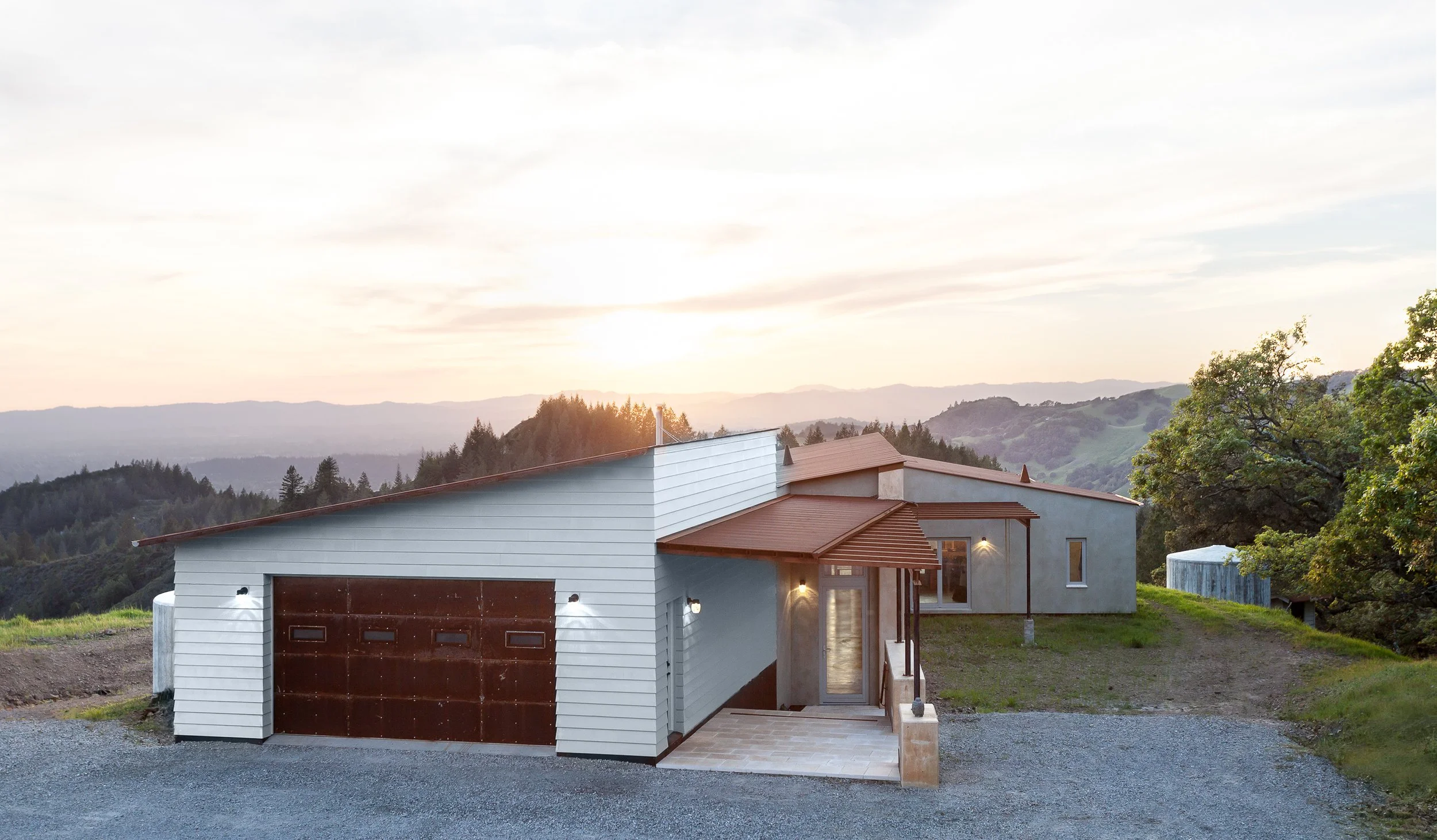 Modern hillside house with garage, surrounded by trees and landscape at sunset.