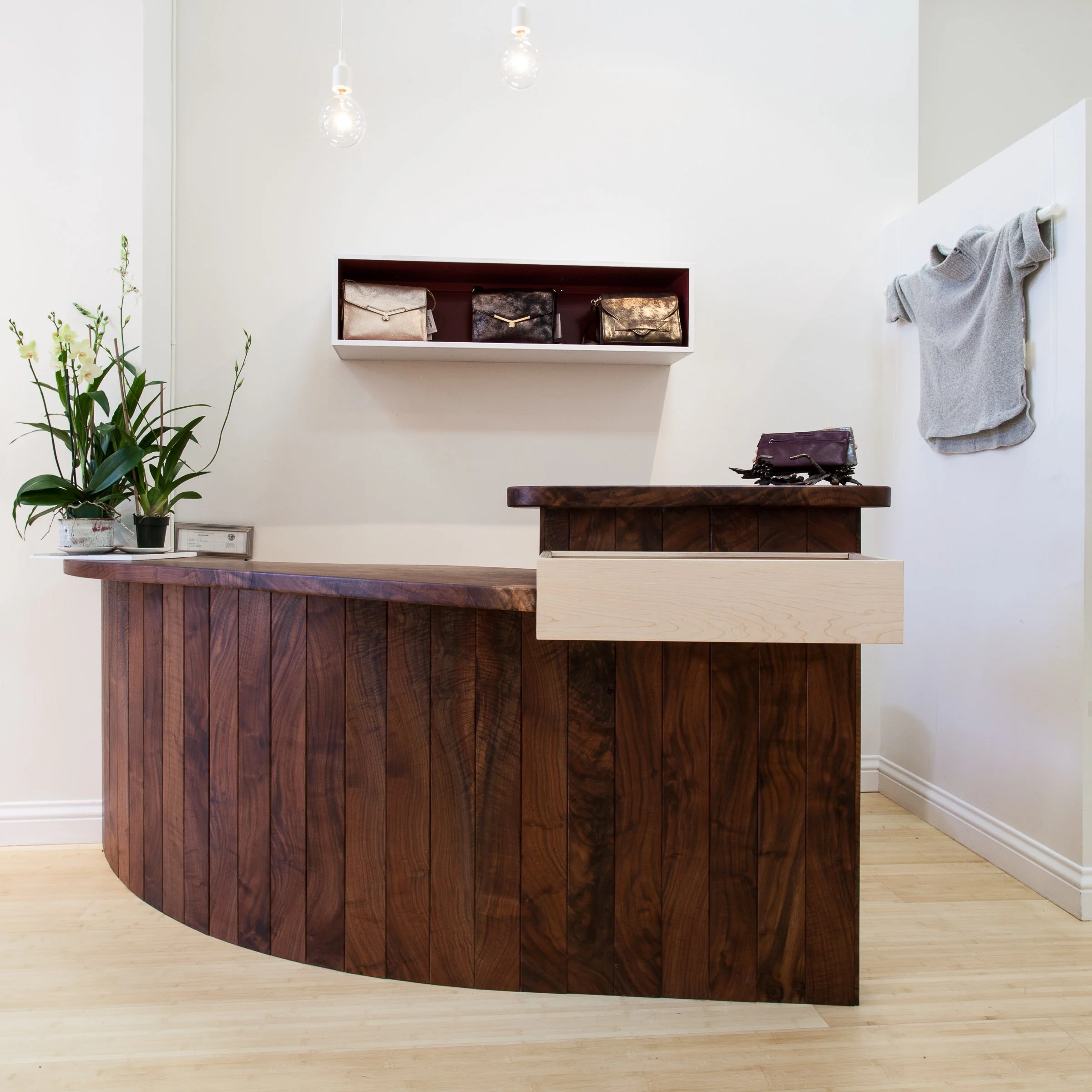 Contemporary wooden reception desk with plants and handbags in a minimalist interior.