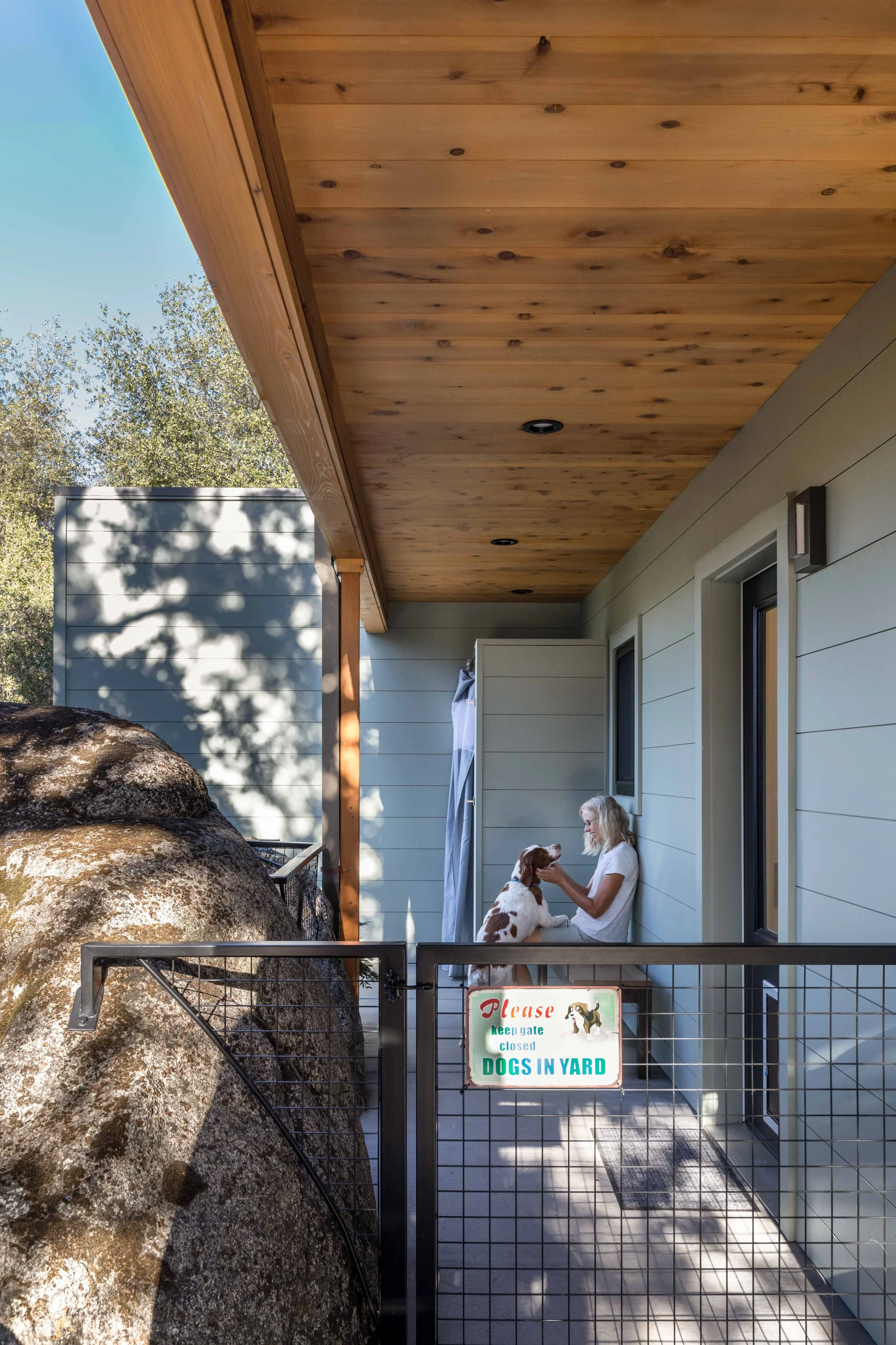 A woman in casual attire sitting on a porch with a dog, beside a metal gate with a sign that reads "Please keep gate closed. Dogs in yard." The porch has a wooden ceiling and siding, and a large rock is visible in the foreground.
