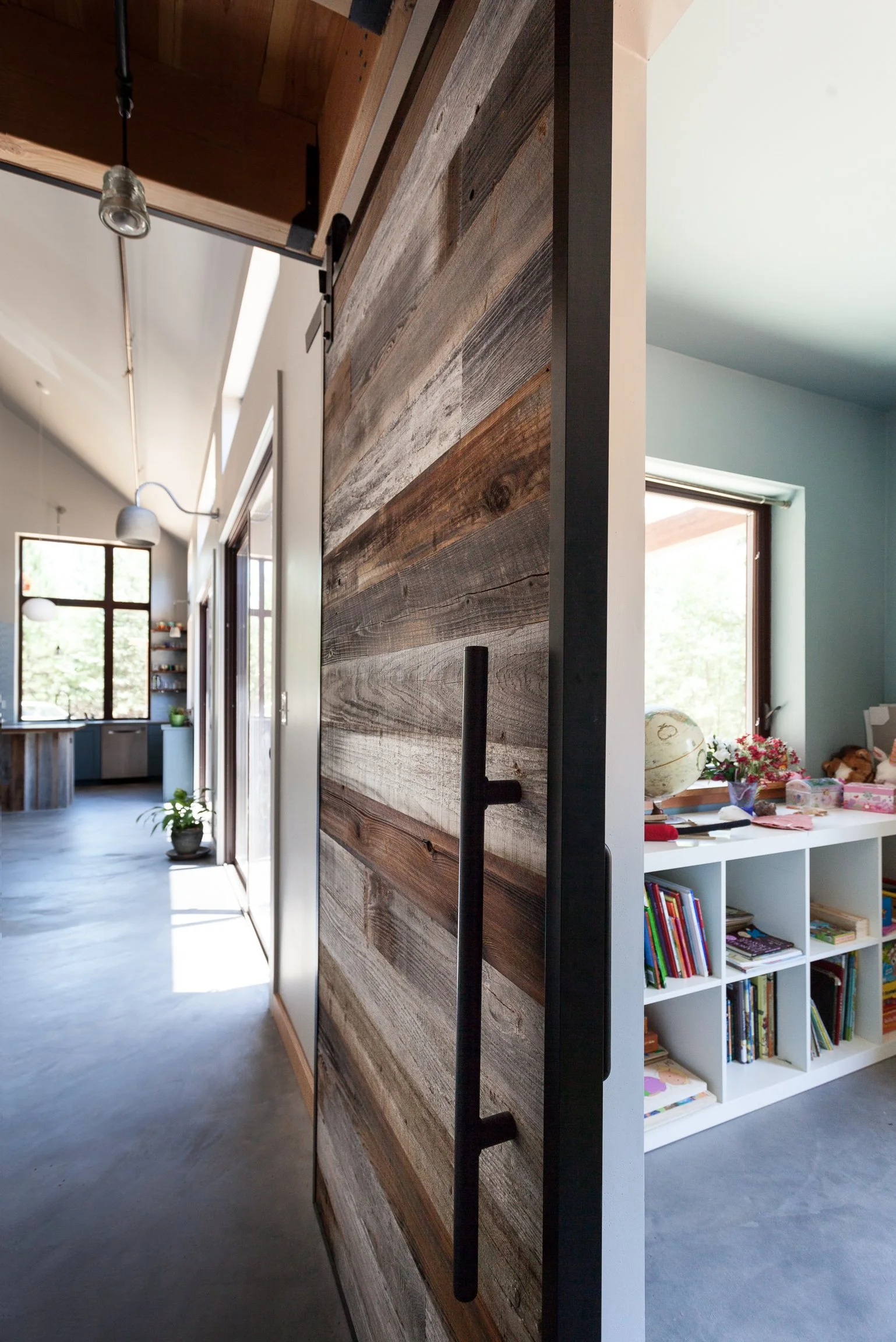 Interior with a sliding barn door made of reclaimed wood, leading to a bright room with a bookshelf filled with books and a globe. The hallway features modern pendant lighting and a plant near the window.
