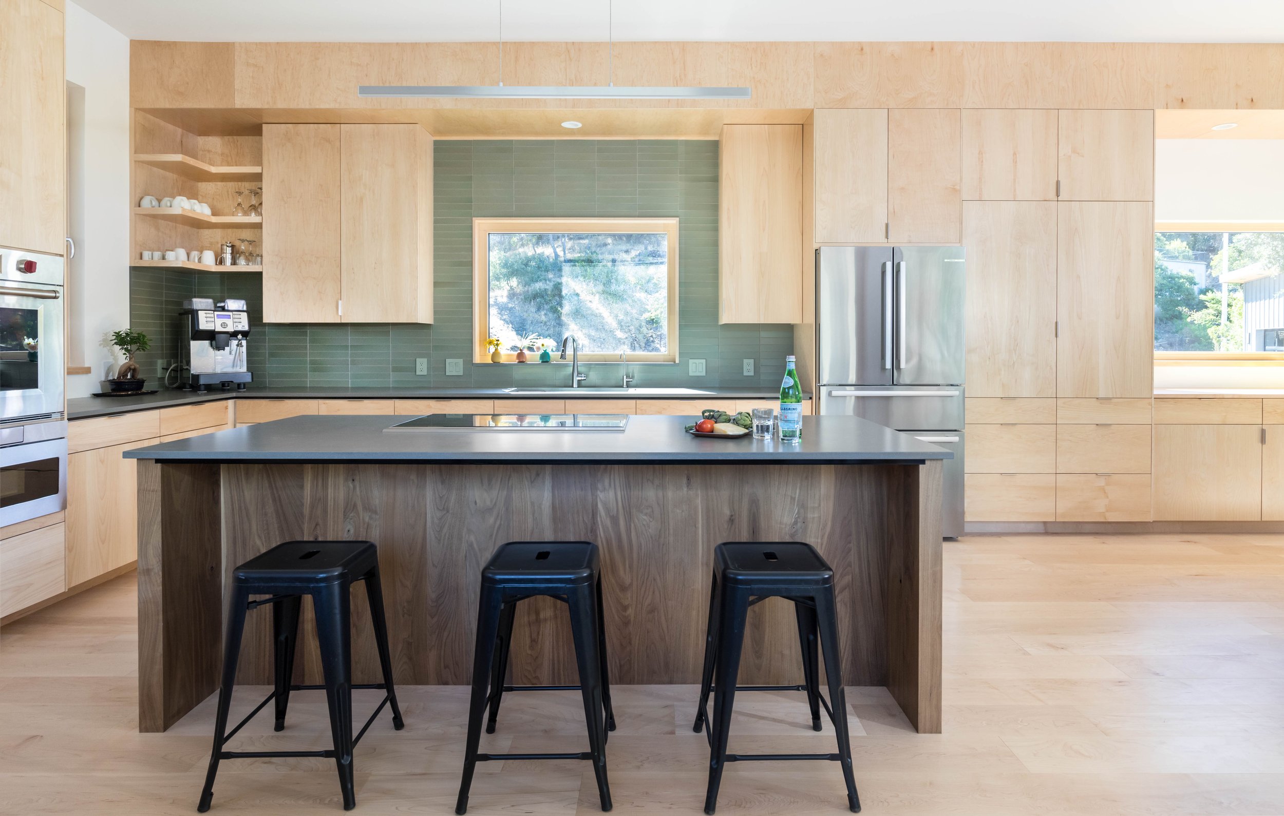 Modern kitchen with wooden cabinets and green backsplash, featuring a central island with three black stools, stainless steel appliances, and a window view of greenery.