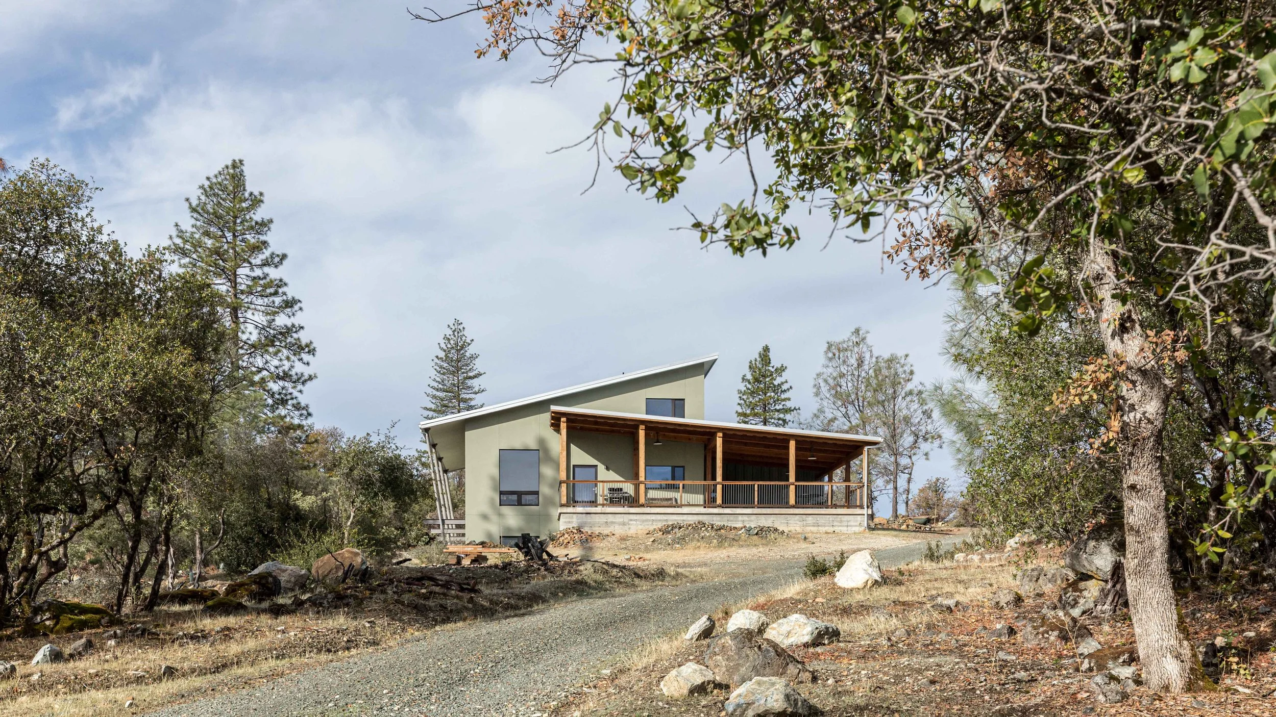 Modern house with a sloped roof surrounded by trees and a gravel path, set in a rural landscape.