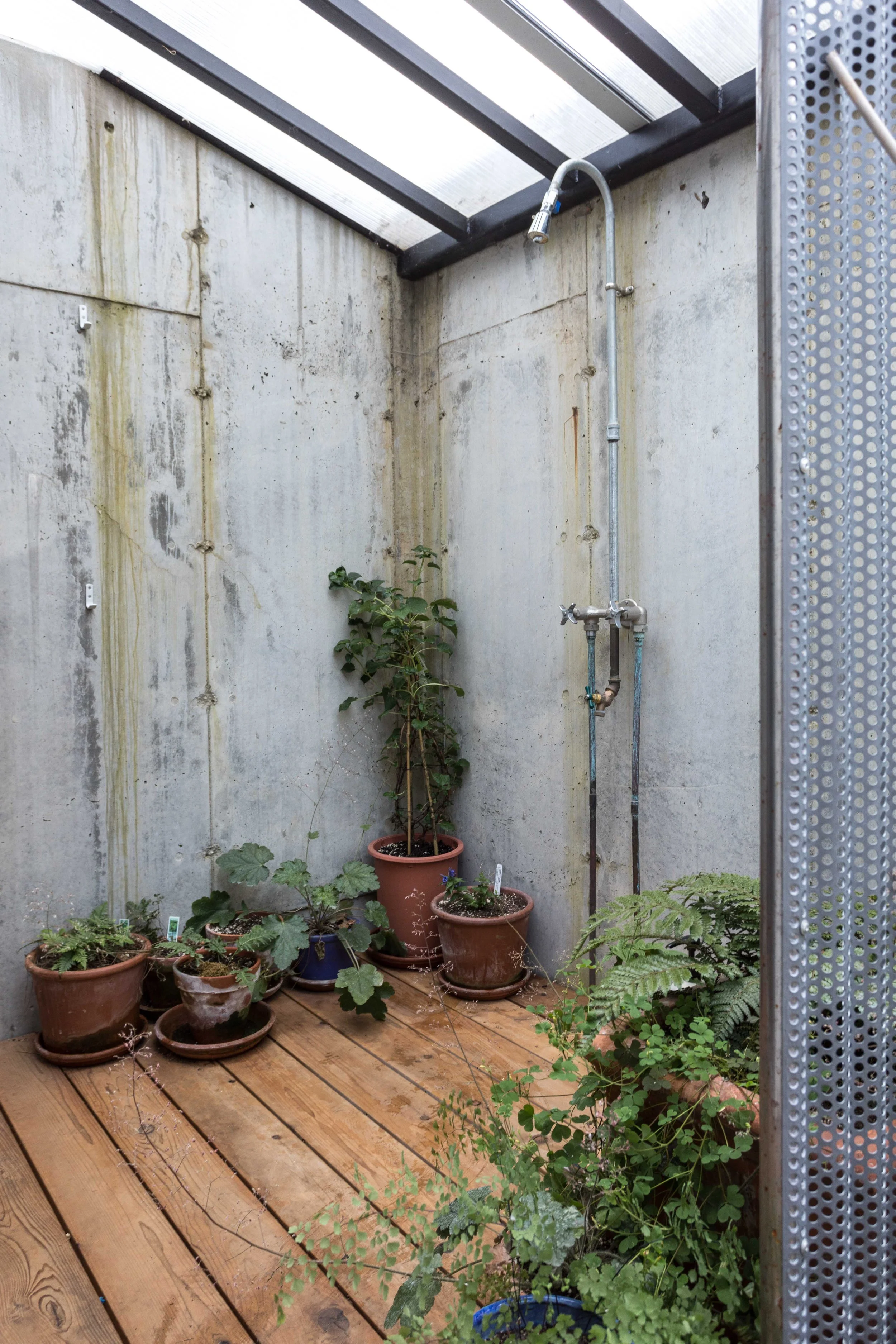 Outdoor shower area with potted plants on a wooden deck, under a clear roof and surrounded by concrete walls.