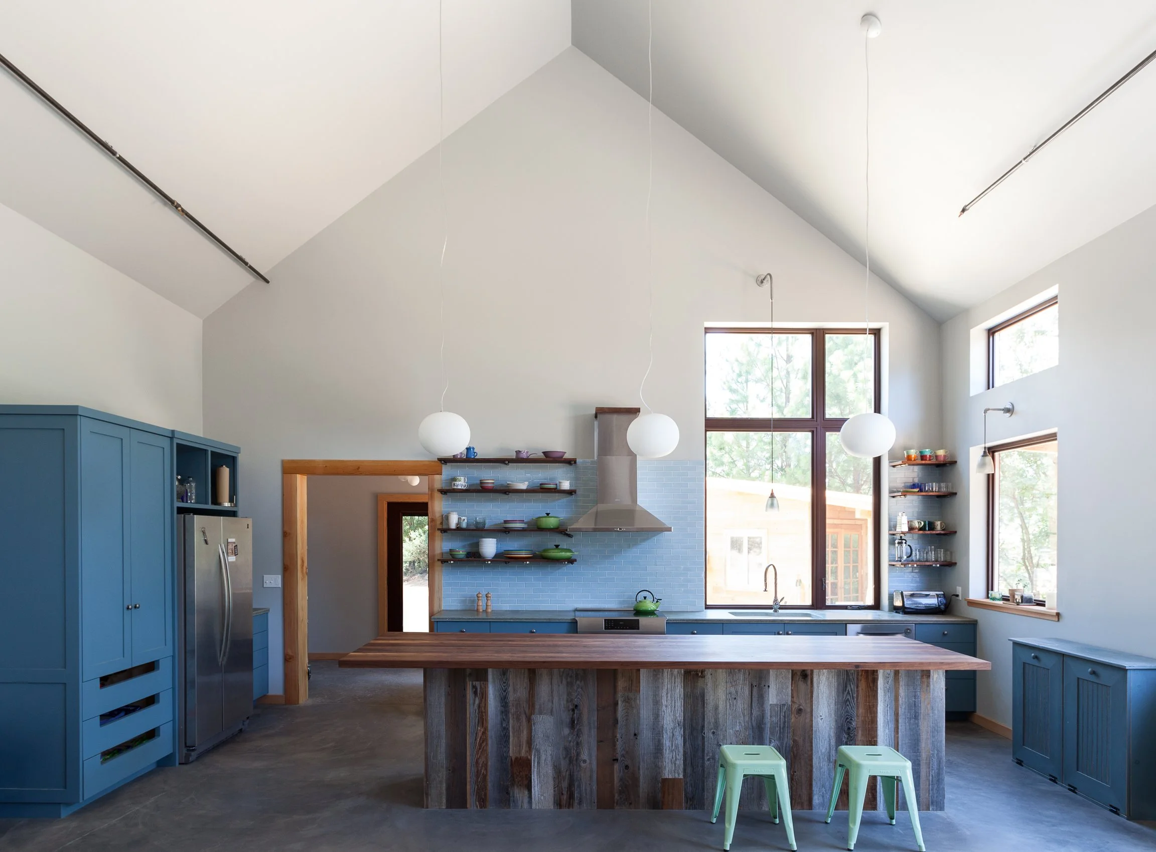 Modern kitchen with blue cabinetry, stainless steel appliances, wooden island, open shelves, large windows, and pendant lighting.