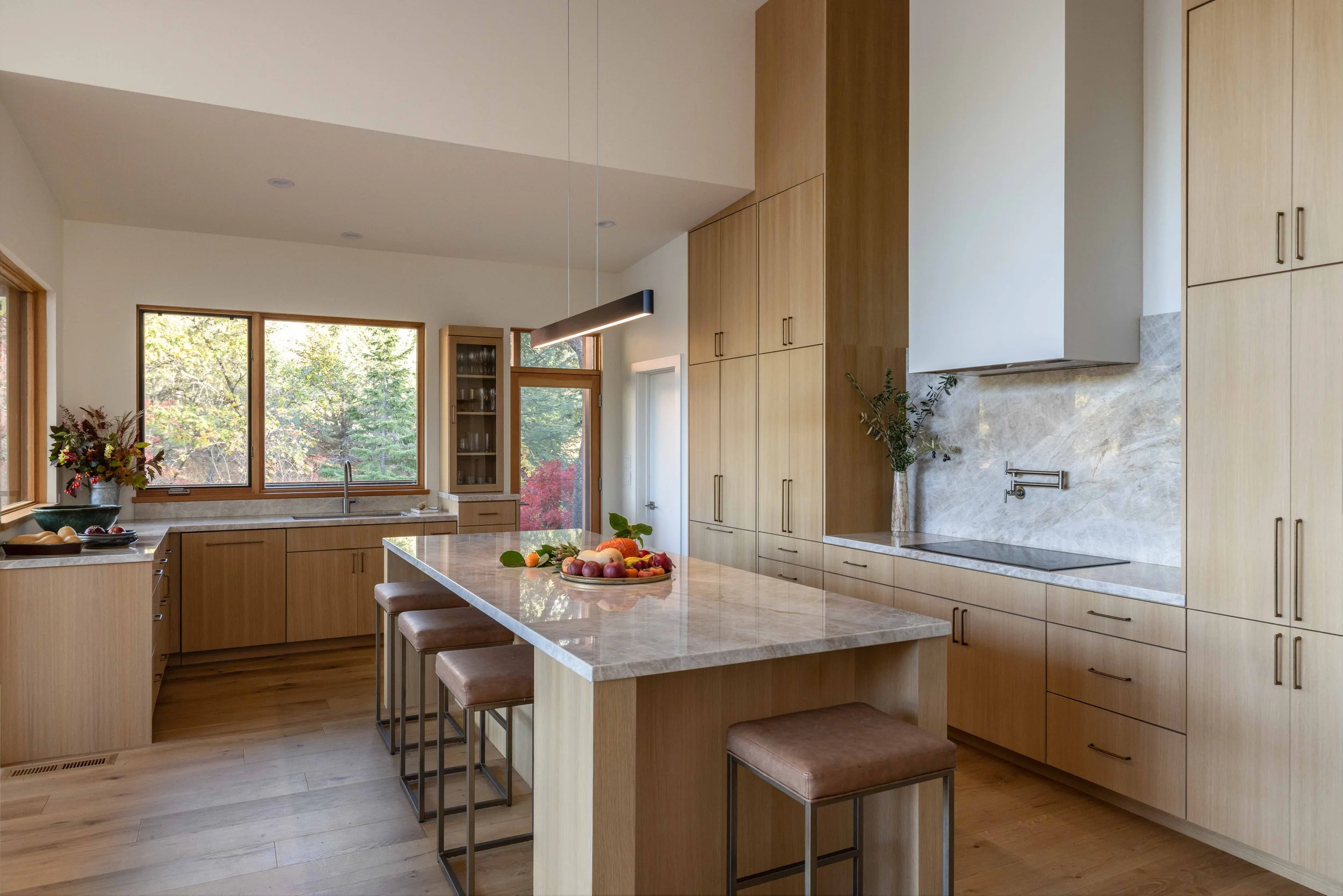 Modern kitchen with wooden cabinets, marble countertops, and an island with stools. Large windows with outdoor view, decorative plants and fruits on the countertop.