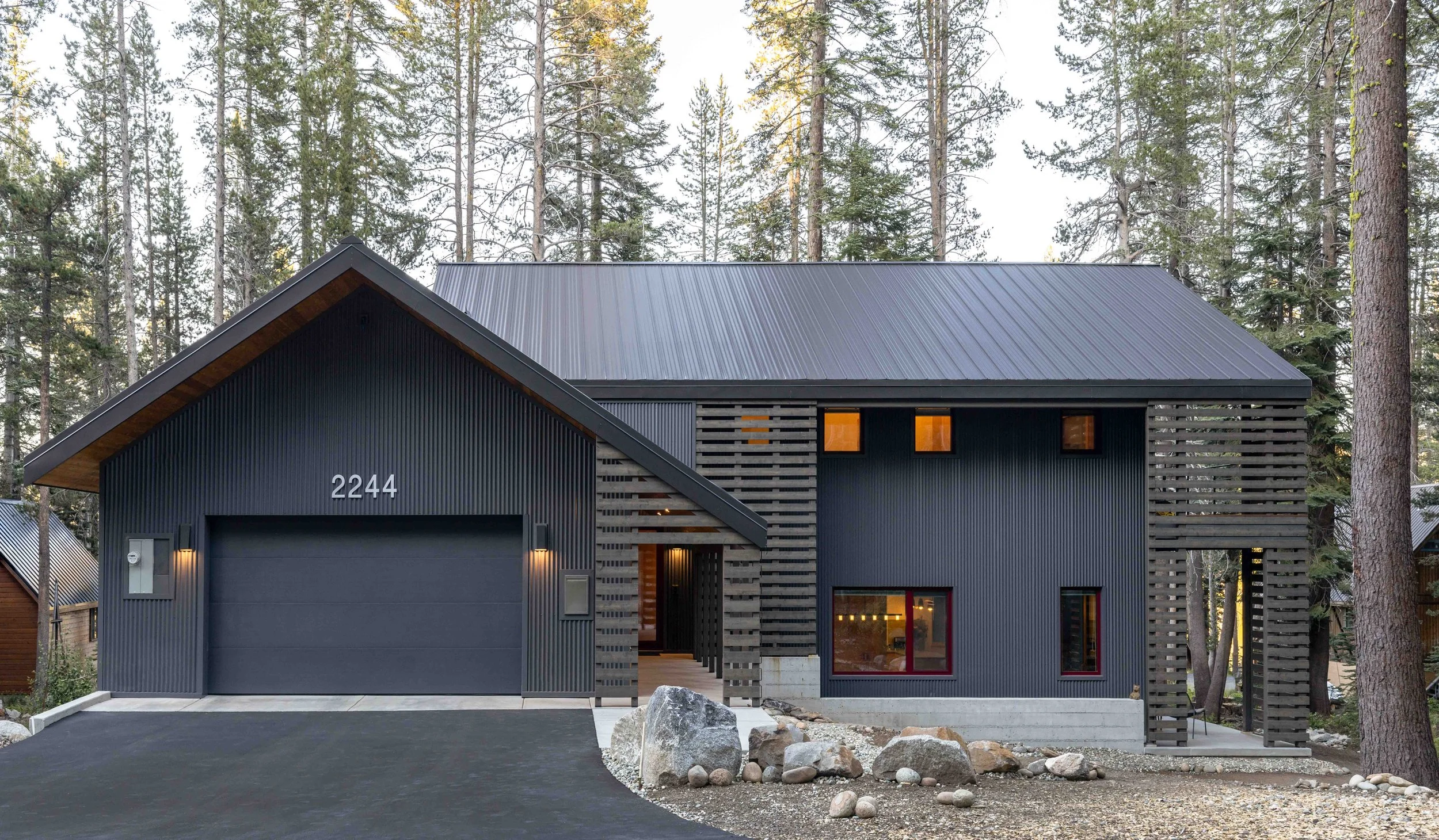 Modern dark gray house with metal roof and wooden accents, surrounded by tall pine trees.