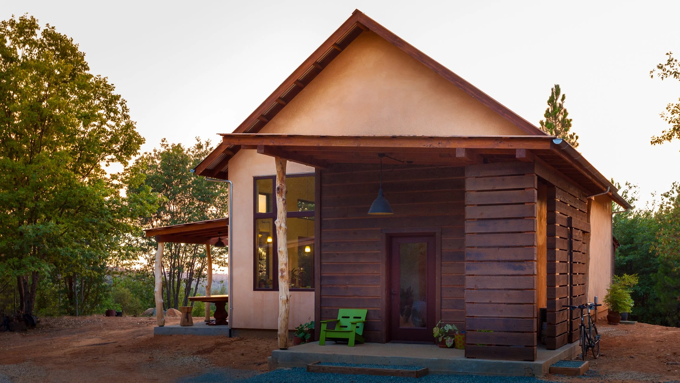 Small house with wood and stucco exterior, surrounded by trees, featuring a front porch with a green chair and a bicycle.