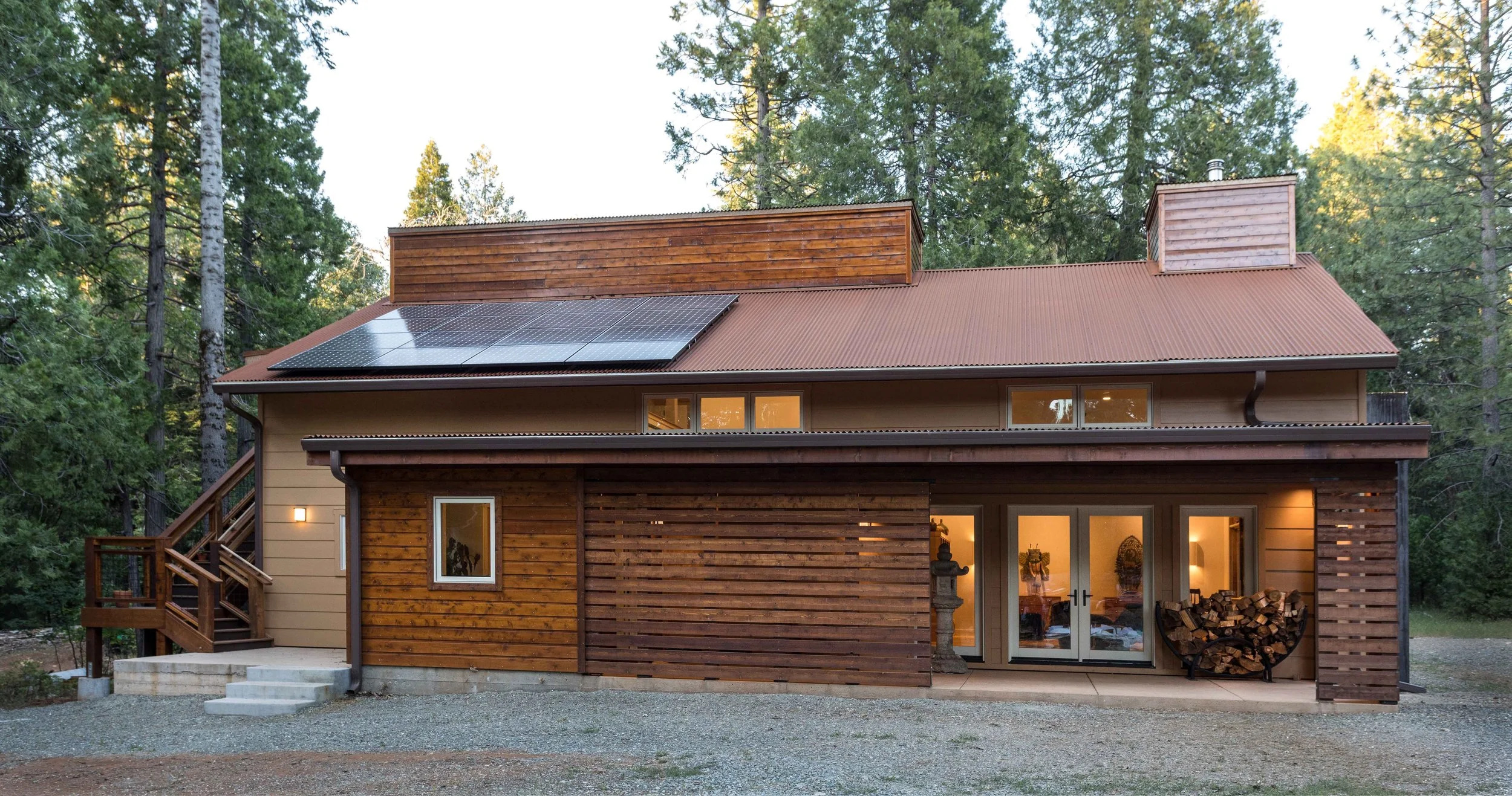 A modern wooden house with solar panels on the roof, surrounded by trees. It features large glass doors and a pile of firewood beside the entrance, with a gravel driveway in front.