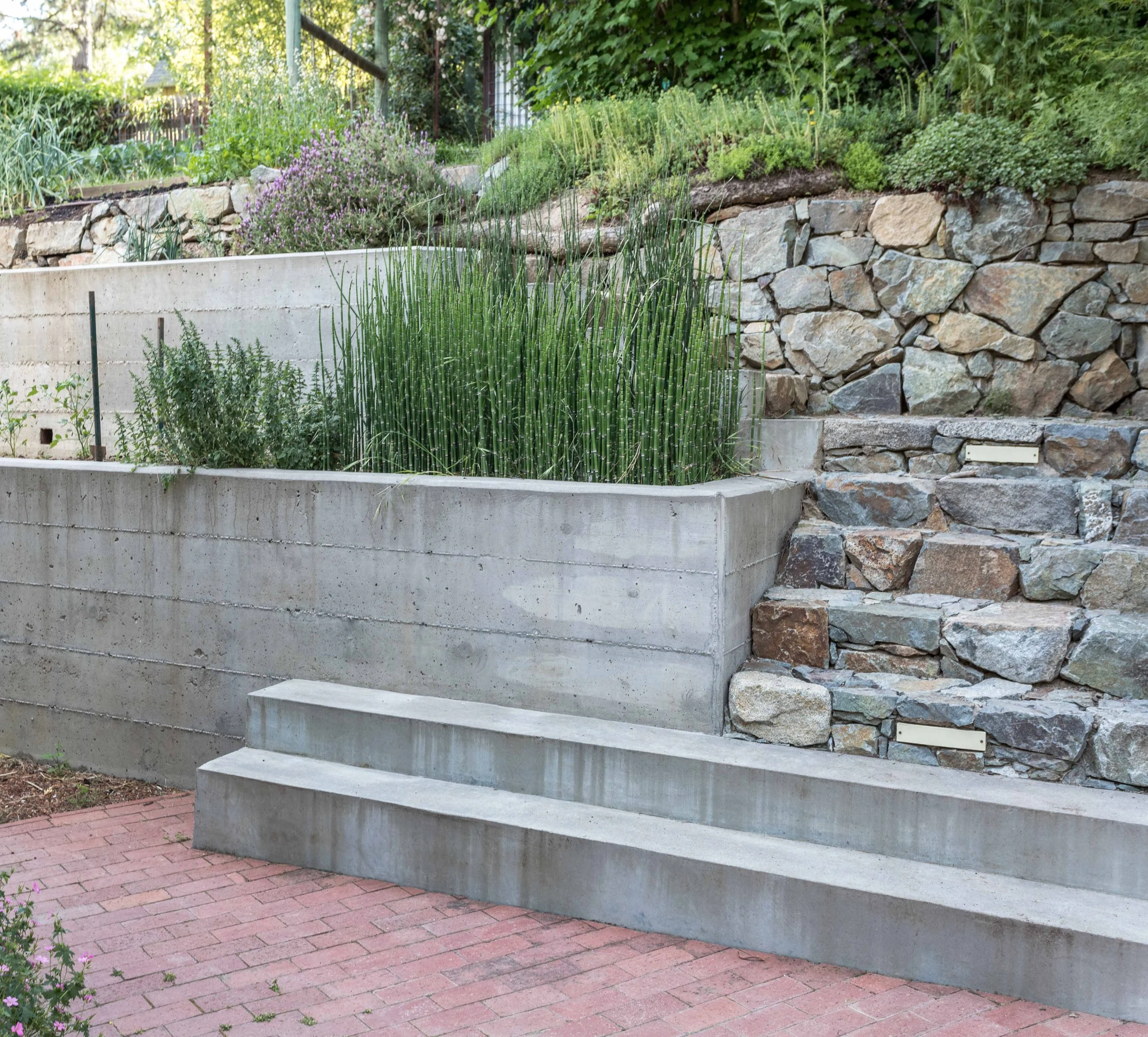 Terraced garden with concrete and stone retaining walls, green plants, brick walkway, and stone steps.