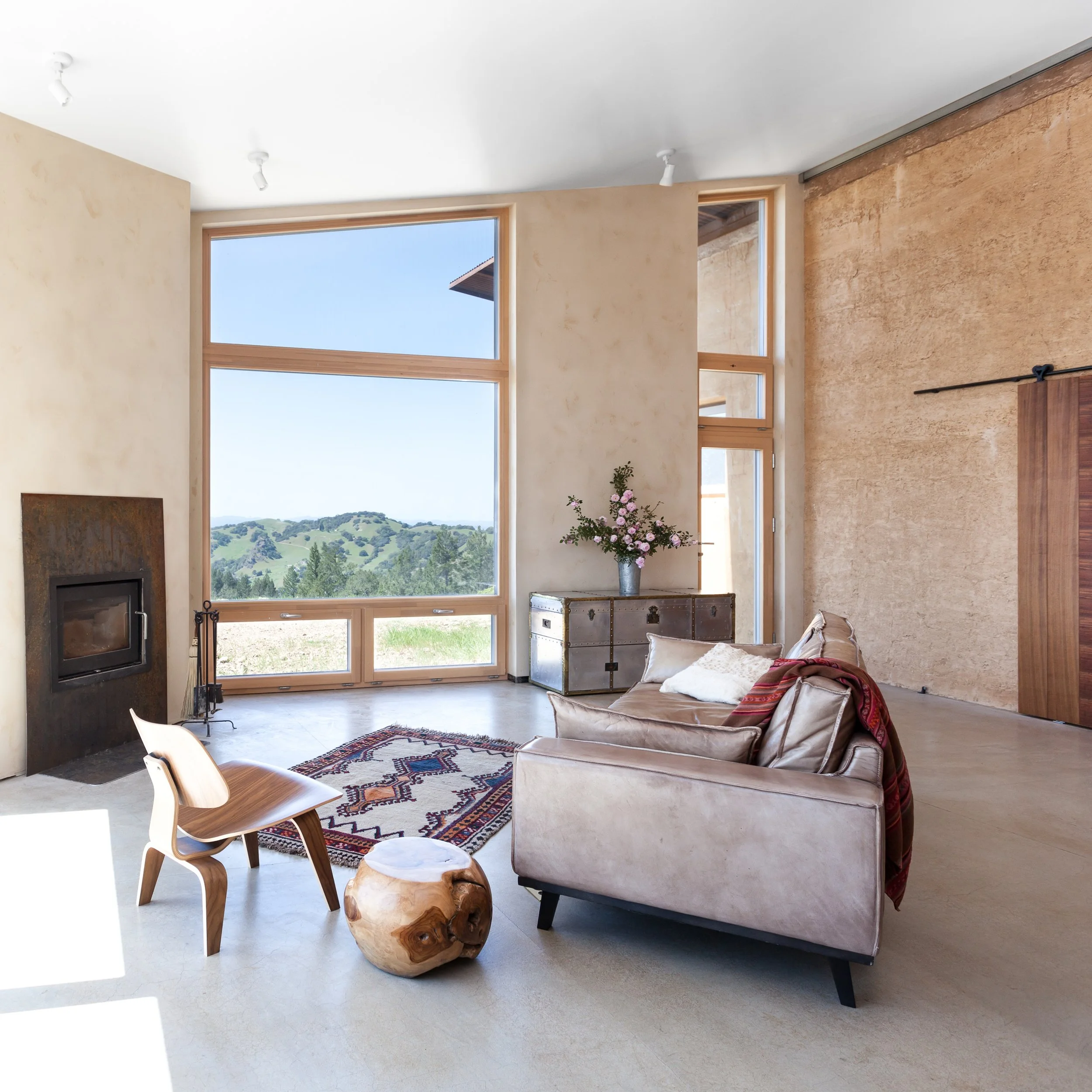 Modern living room with large windows, a landscape view, a leather sofa, wooden chair, and rustic decorations. A colorful rug, fireplace, and decorative vases add to the cozy ambiance.