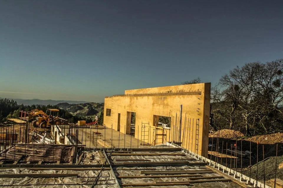 Construction site with partially built yellow wall, surrounded by reinforcing bars and construction equipment, set against a hilly landscape and clear sky.