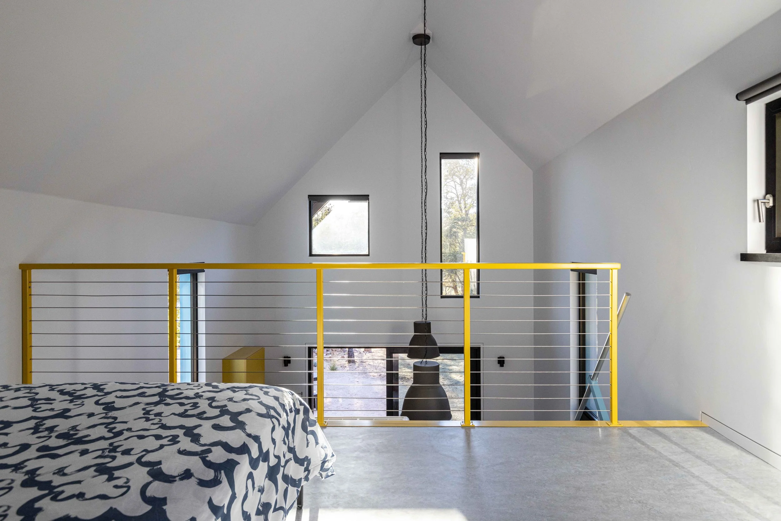 Modern loft with yellow railing, abstract patterned bedspread, high ceiling, and large pendant light. Features two windows with outdoor view.