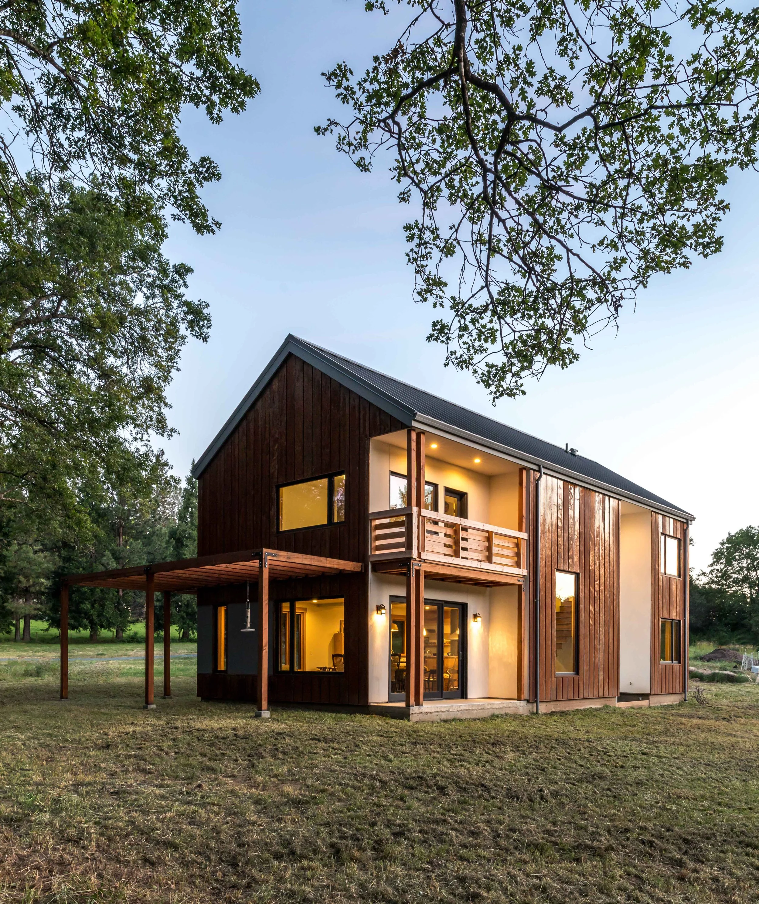 Modern two-story house with wooden exterior, large windows, and a small balcony, surrounded by trees and grass, during dusk.