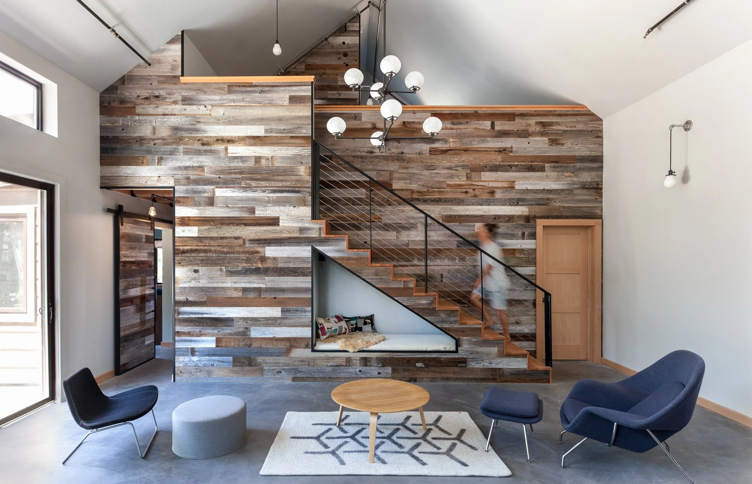 Modern interior with a wooden accent wall, staircase, and blue armchairs. A person walks up the stairs. A round wooden table and pouf are on a patterned rug beneath a contemporary chandelier.