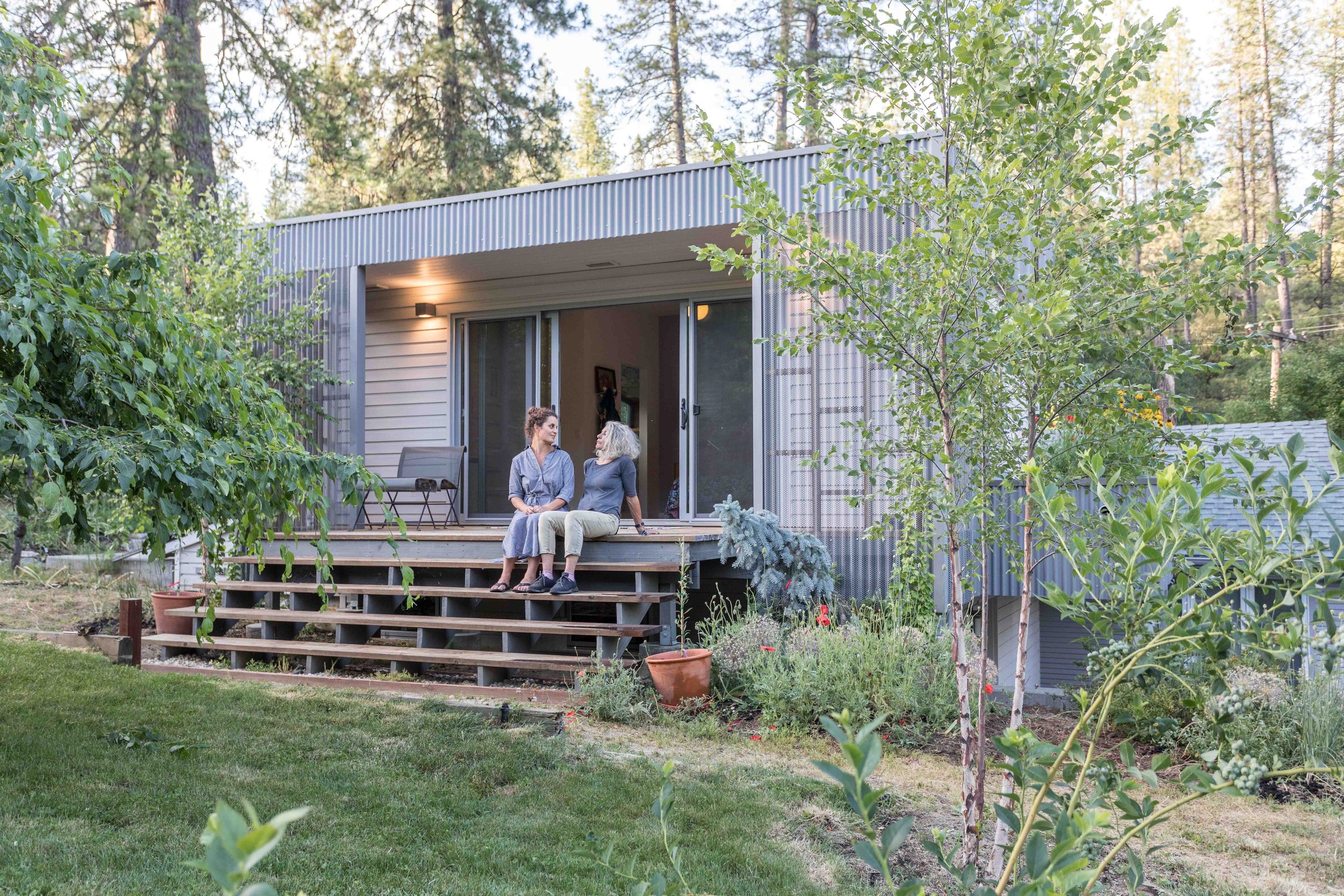 Two people sitting on the porch steps of a small modern house surrounded by trees and garden plants.