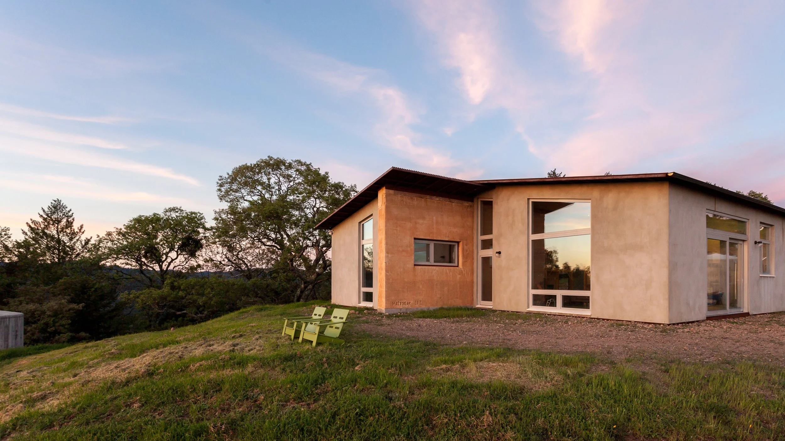 Modern eco-friendly house on a grassy hill with large windows and a minimalist design, surrounded by trees under a clear sky.