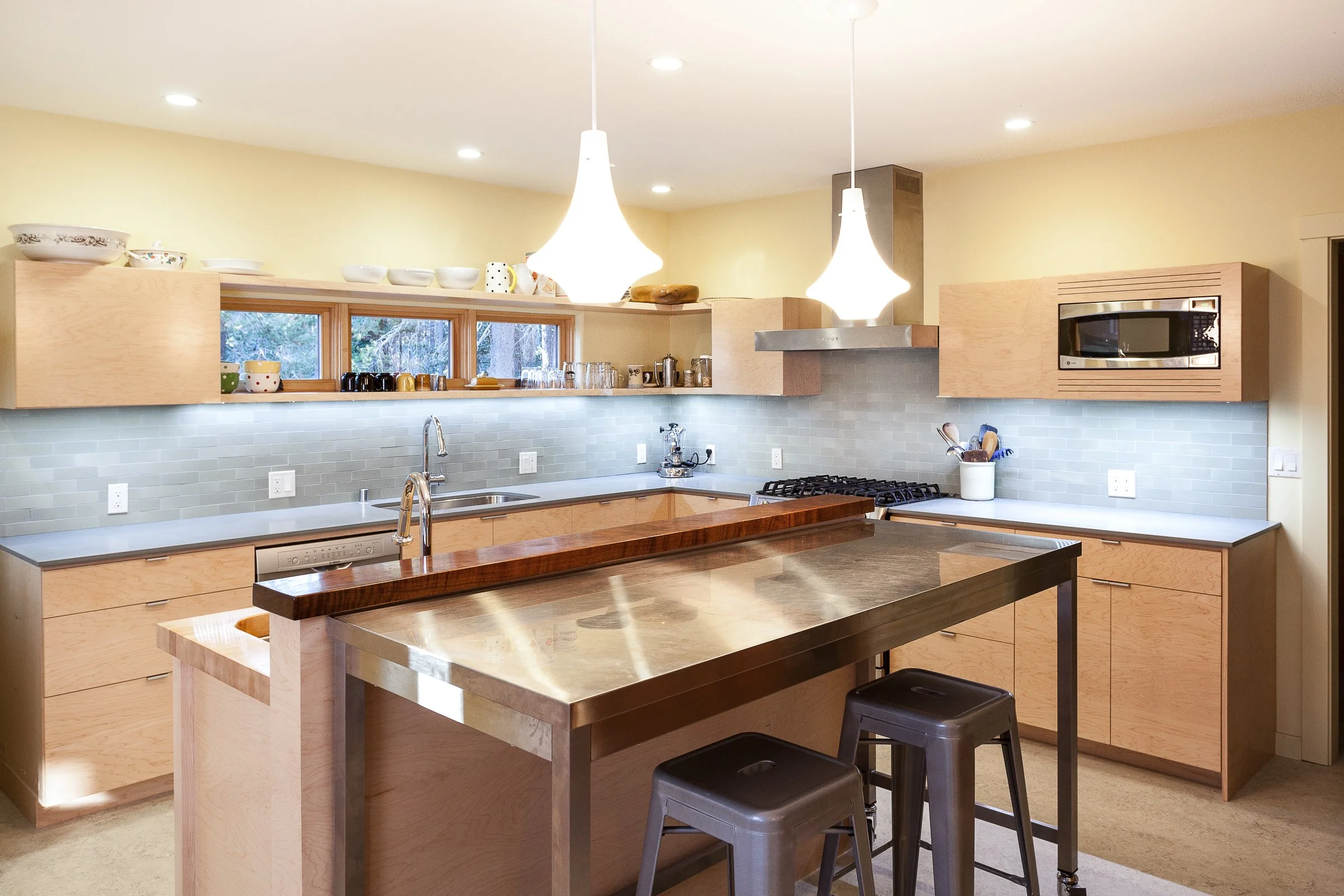 Modern kitchen with light wood cabinets, stainless steel appliances, and an island. Features a double sink, gas stove, microwave, recessed lighting, and pendant lights. Gray subway tile backsplash and bar seating with metal stools.