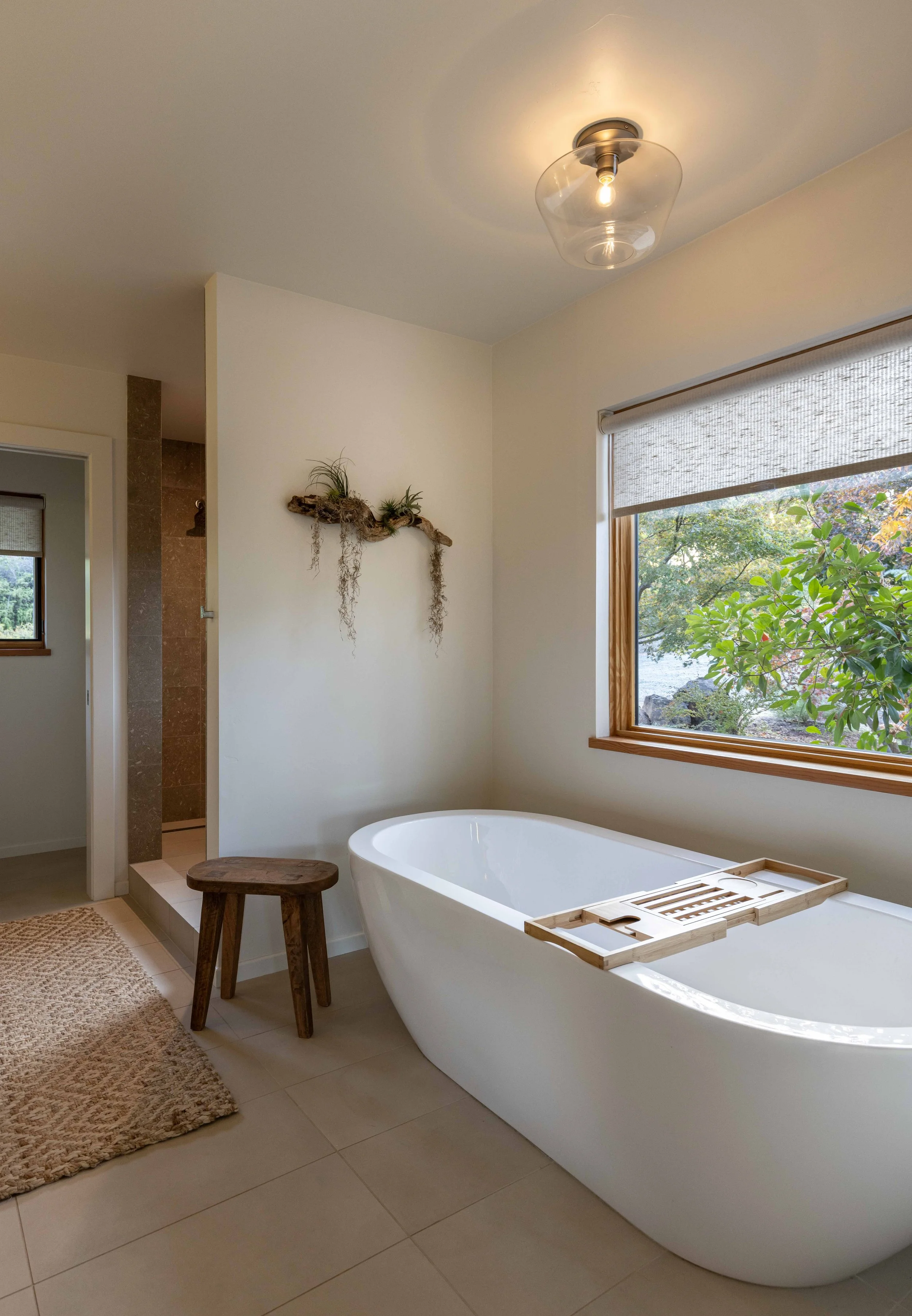 Modern bathroom with freestanding bathtub, wooden stool, wall-mounted decorative shelf, large window with outdoor view, beige tiles, and a ceiling light fixture.
