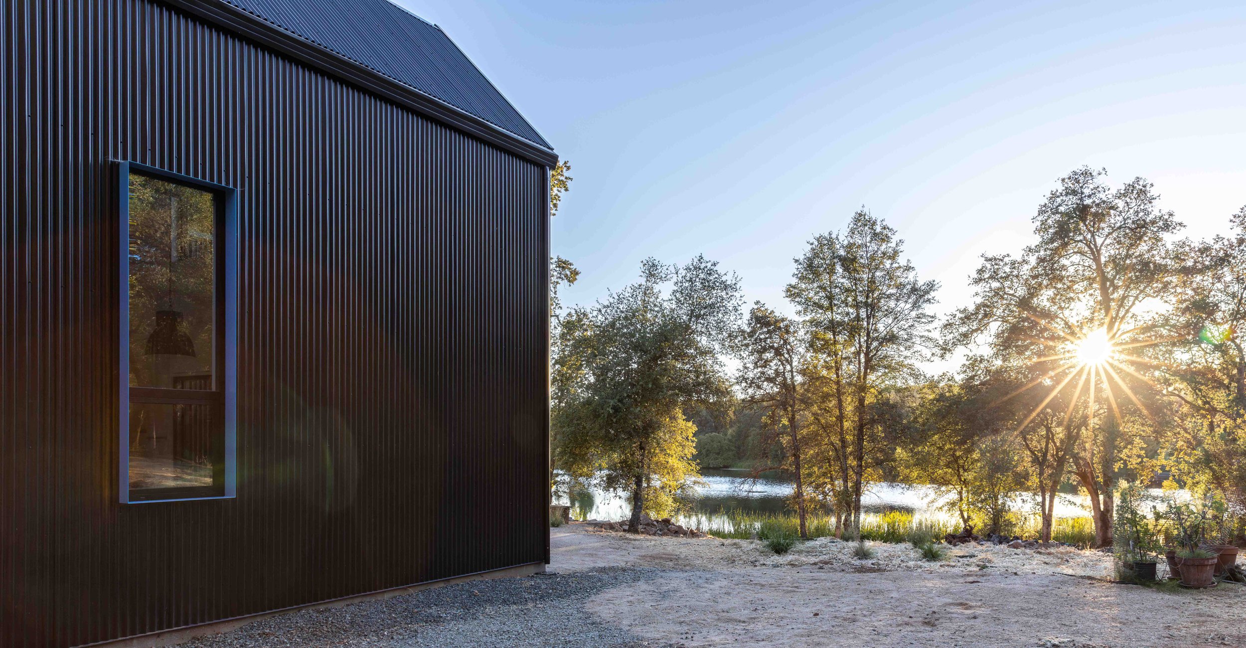 Modern building exterior with corrugated metal siding, surrounded by trees and a lake, with sun shining through trees.