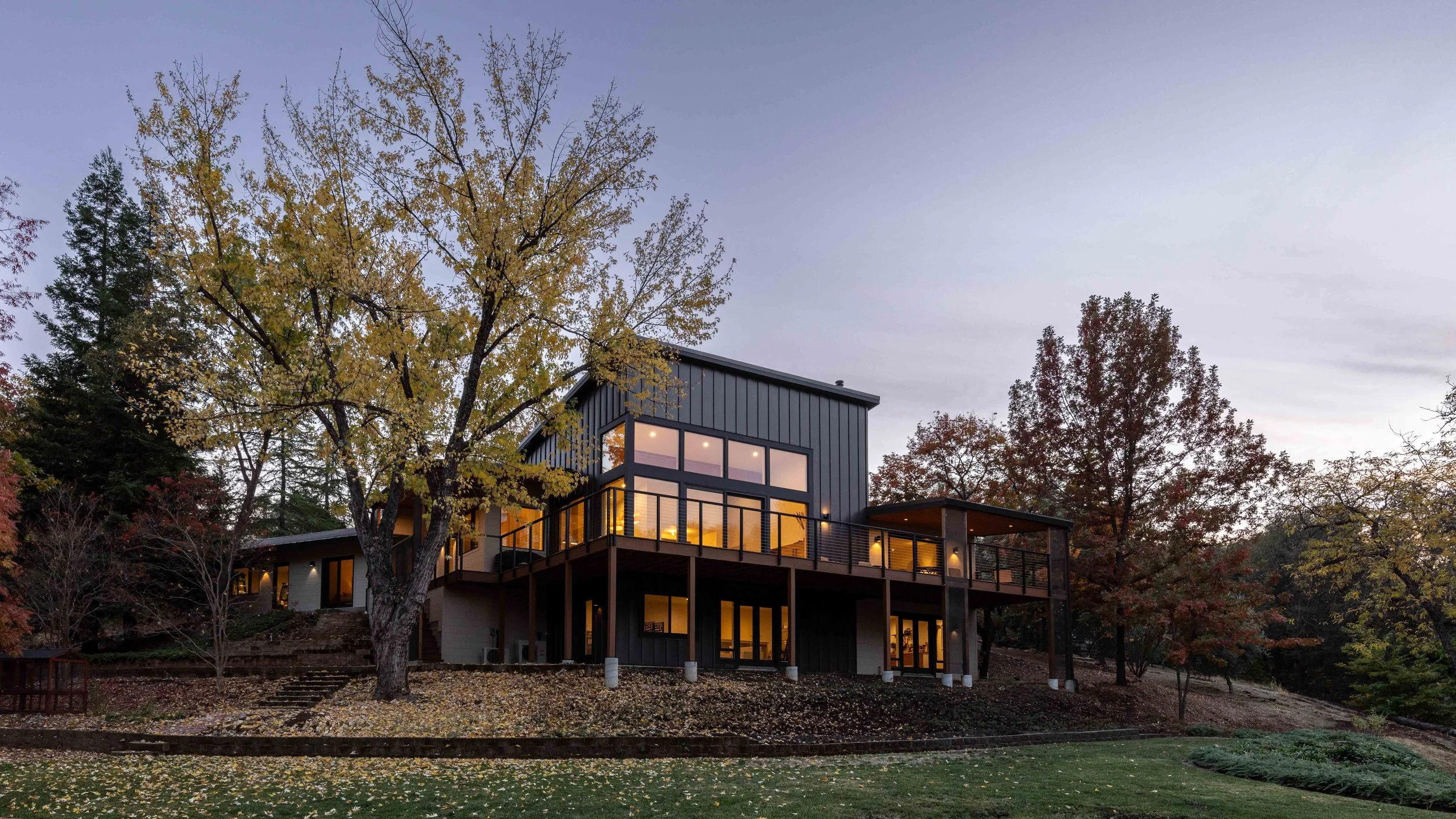 Modern two-story house with large windows and exterior lighting, surrounded by autumn trees and fallen leaves, during twilight.
