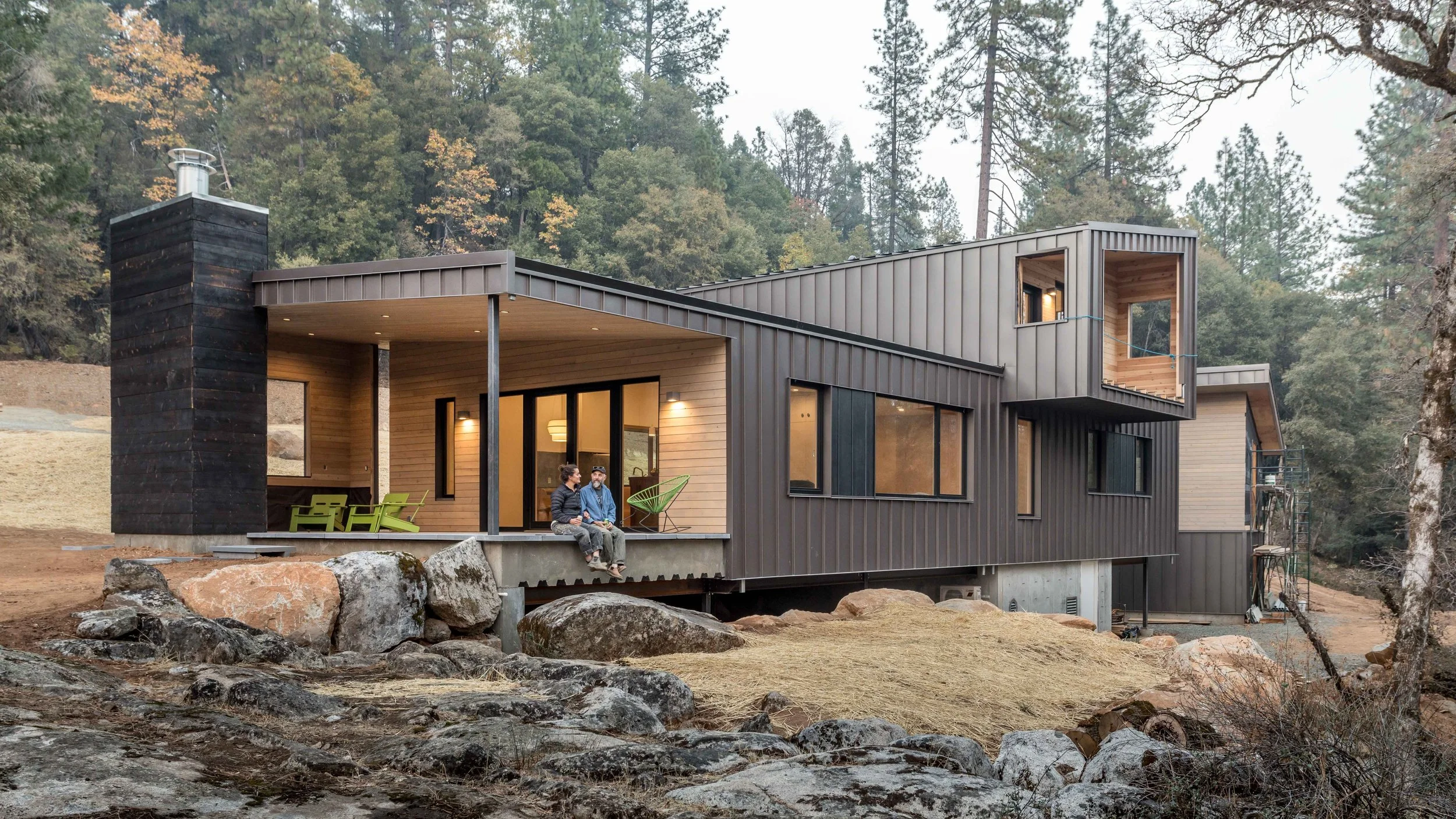 Modern house with large windows and wooden exterior in a forest setting, two people sitting on the porch.