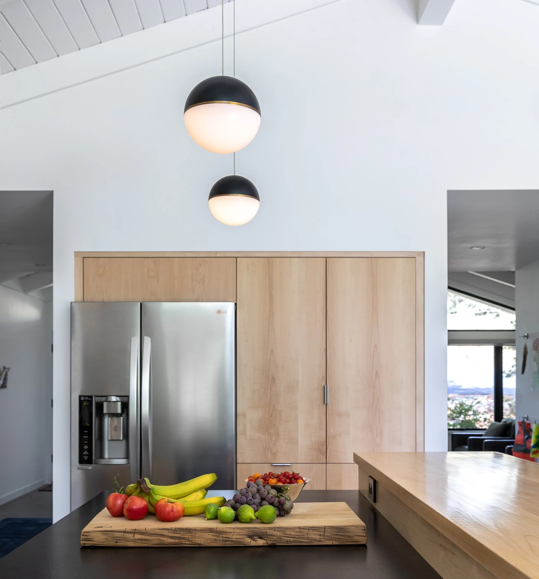 Modern kitchen with stainless steel fridge, wooden cabinets, and pendant lights. A cutting board with bananas, tomatoes, grapes, and limes is on the countertop.
