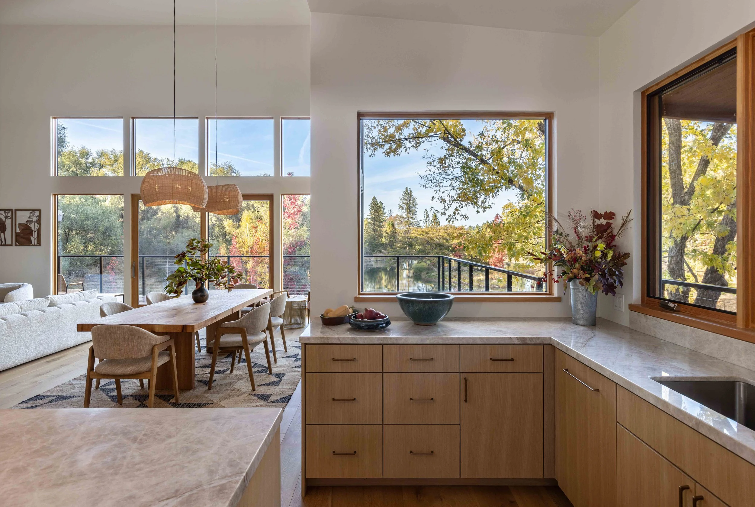 Modern open-plan kitchen and dining area with large windows showing scenic outdoor views, wooden cabinetry, a marble countertop, and a dining table with chairs. Natural light highlights design elements, including pendant lights and decorative plants.