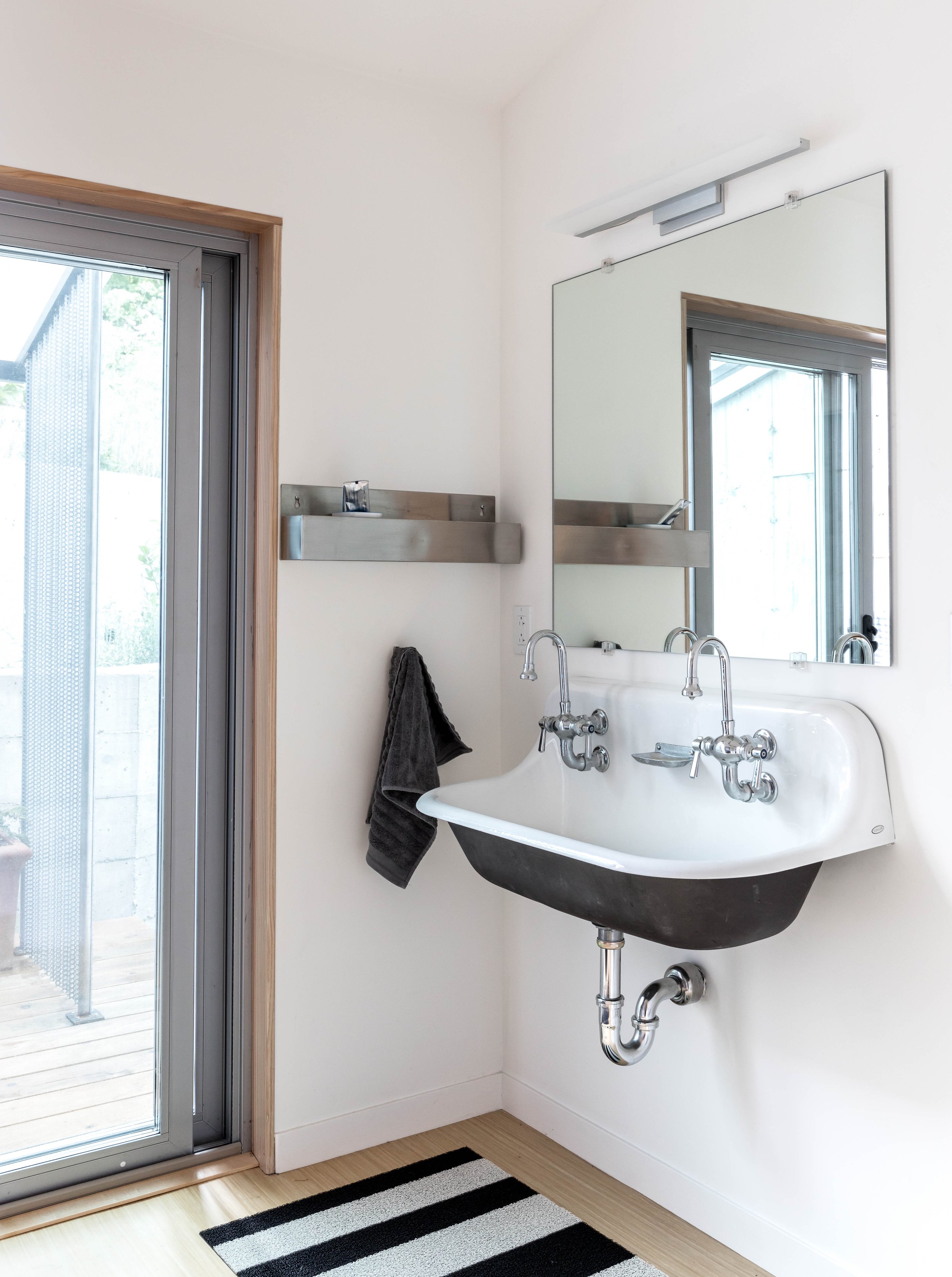 Modern bathroom with a double sink, large mirror, and wall-mounted metal shelf. A black towel hangs beside the sink. A sliding glass door leads to a wooden deck. A black and white striped rug is on the floor.