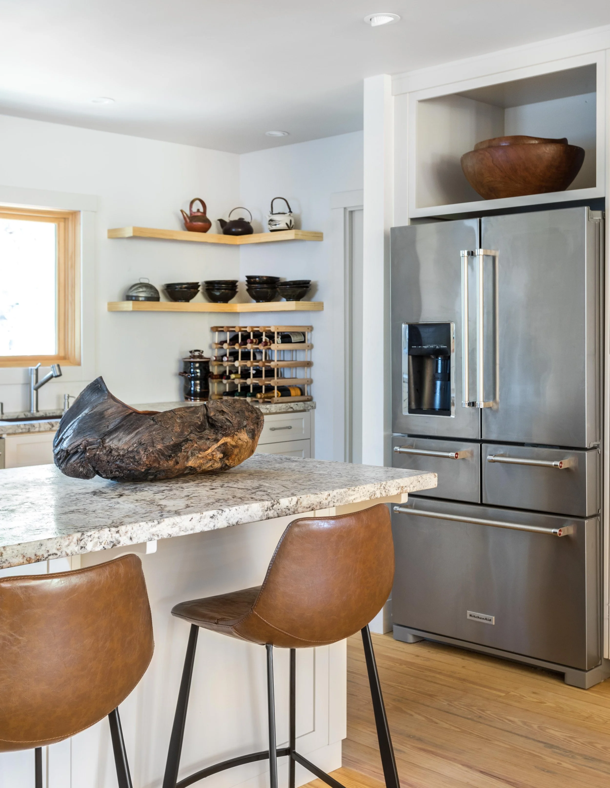 Modern kitchen with granite island, leather stools, stainless steel refrigerator, wooden decor, and open shelves with teapots and bowls.