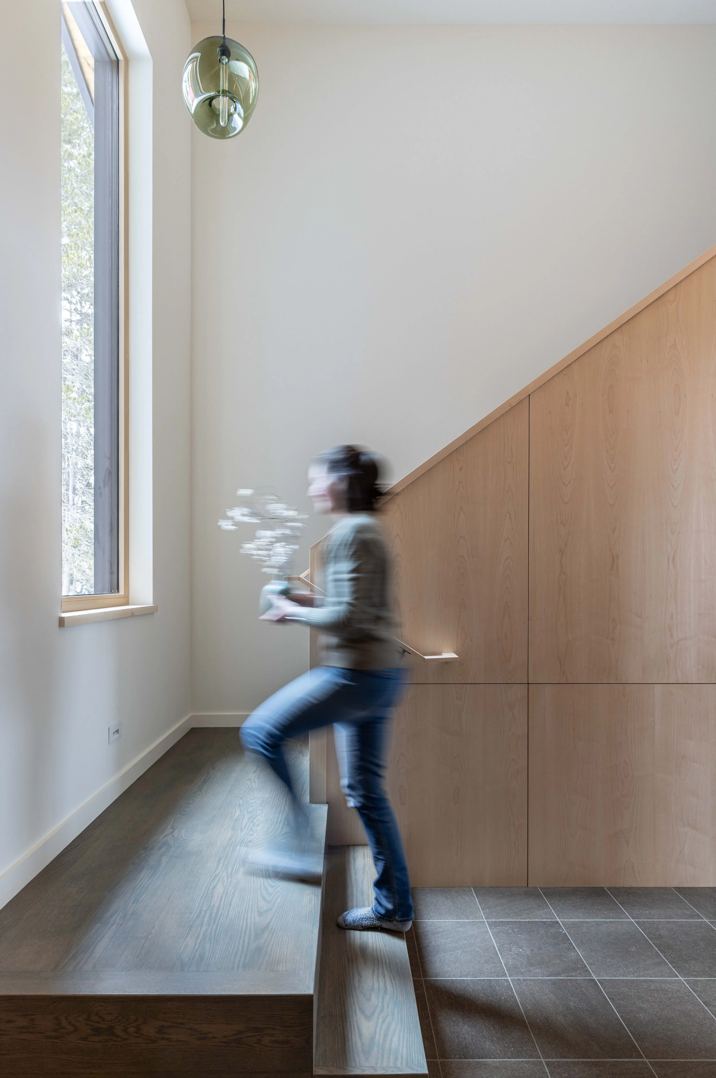 Person walking up stairs indoors holding a vase with flowers, near a large window, with a modern pendant light above and wooden paneling on the side.