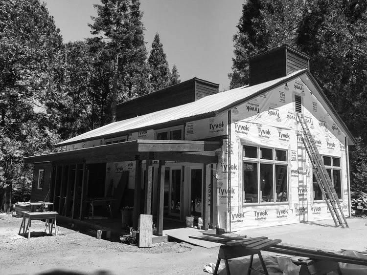 House under construction with Tyvek wrap, surrounded by trees, ladder leaning against the front, building materials scattered around.