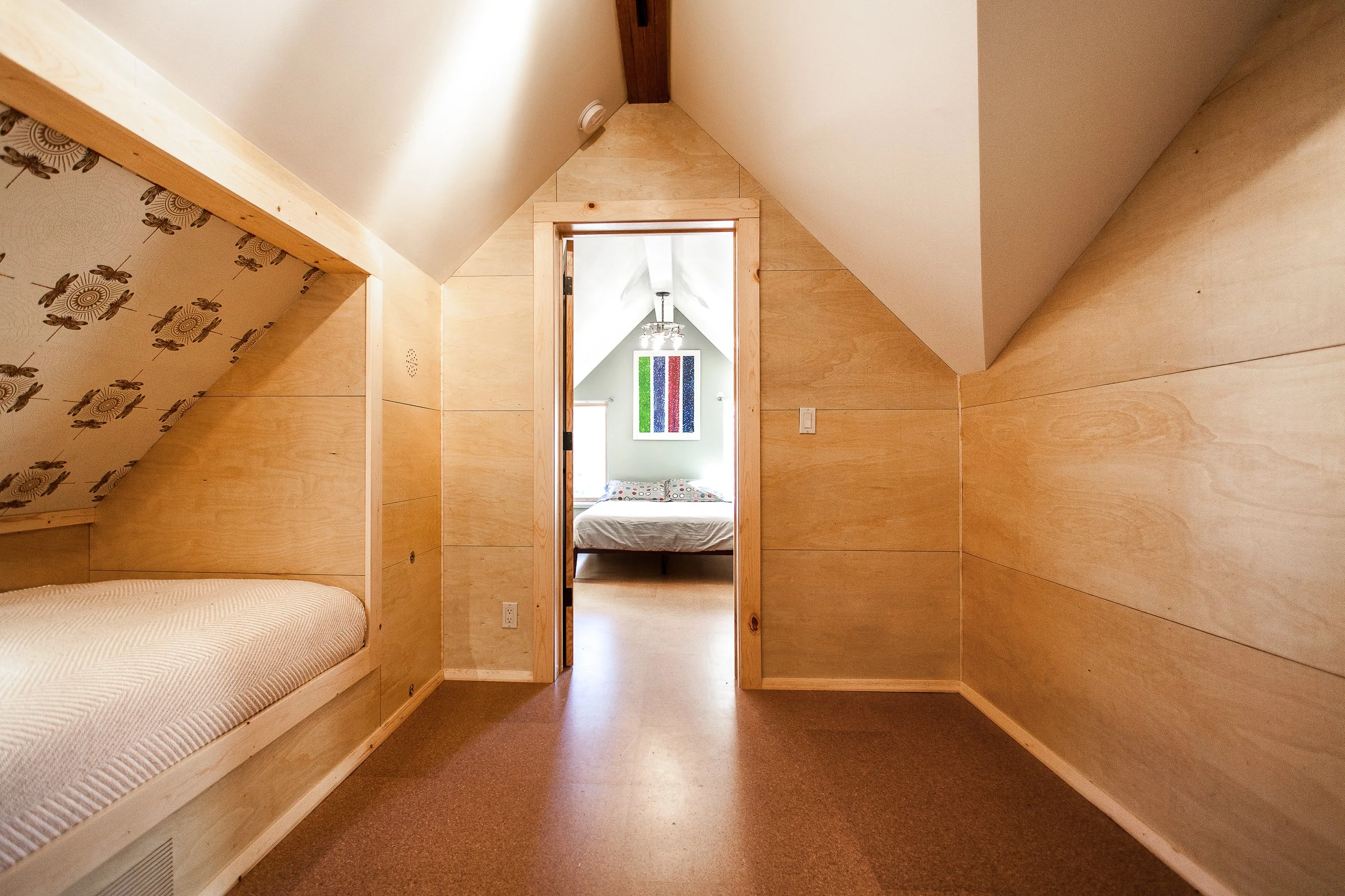 Small attic bedroom with wooden walls, a bunk bed on the left, and a view into another room with a larger bed. Minimalist design and sloped ceiling.