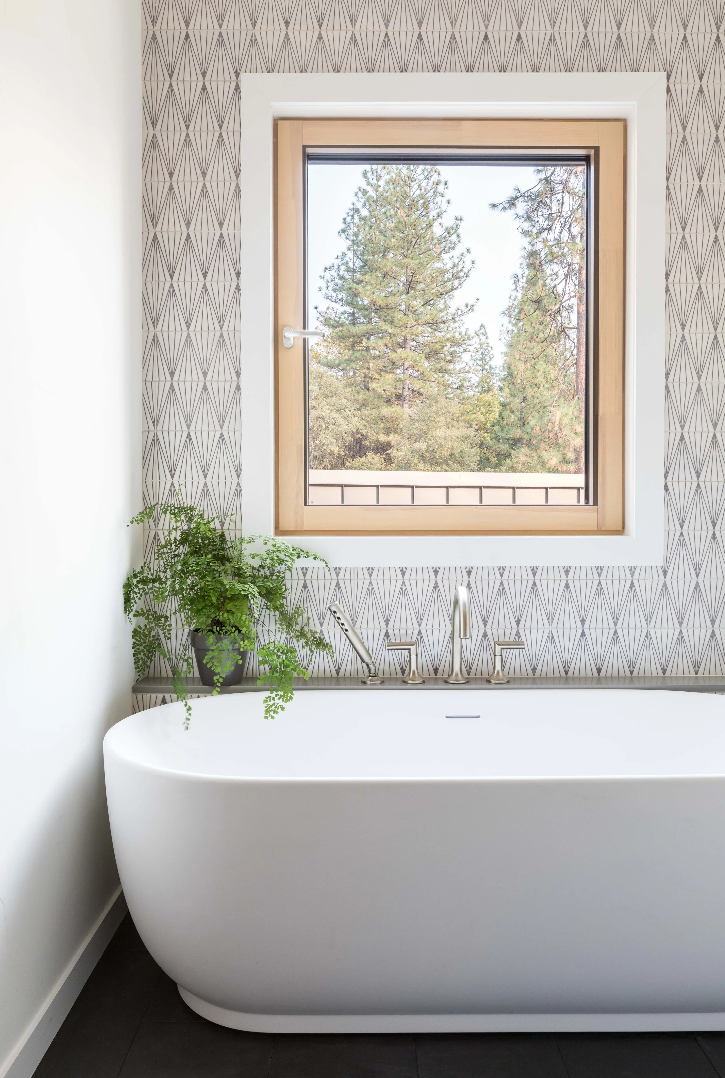 Modern bathroom with a white freestanding bathtub, geometric wallpaper, potted plant, and a window with a view of trees.