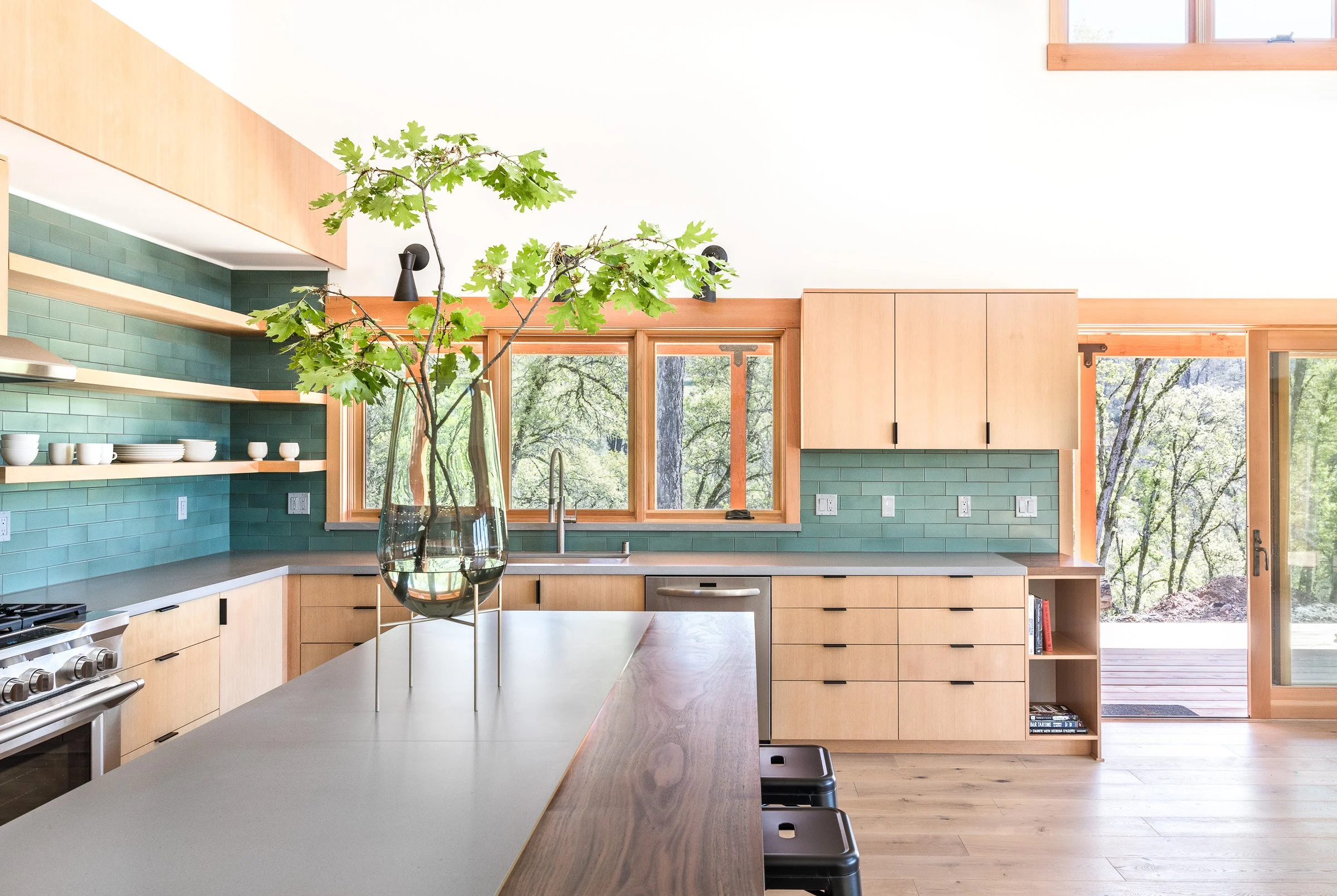 Modern kitchen with light wood cabinets, green tiled backsplash, and large windows; a vase with green branches on the island.
