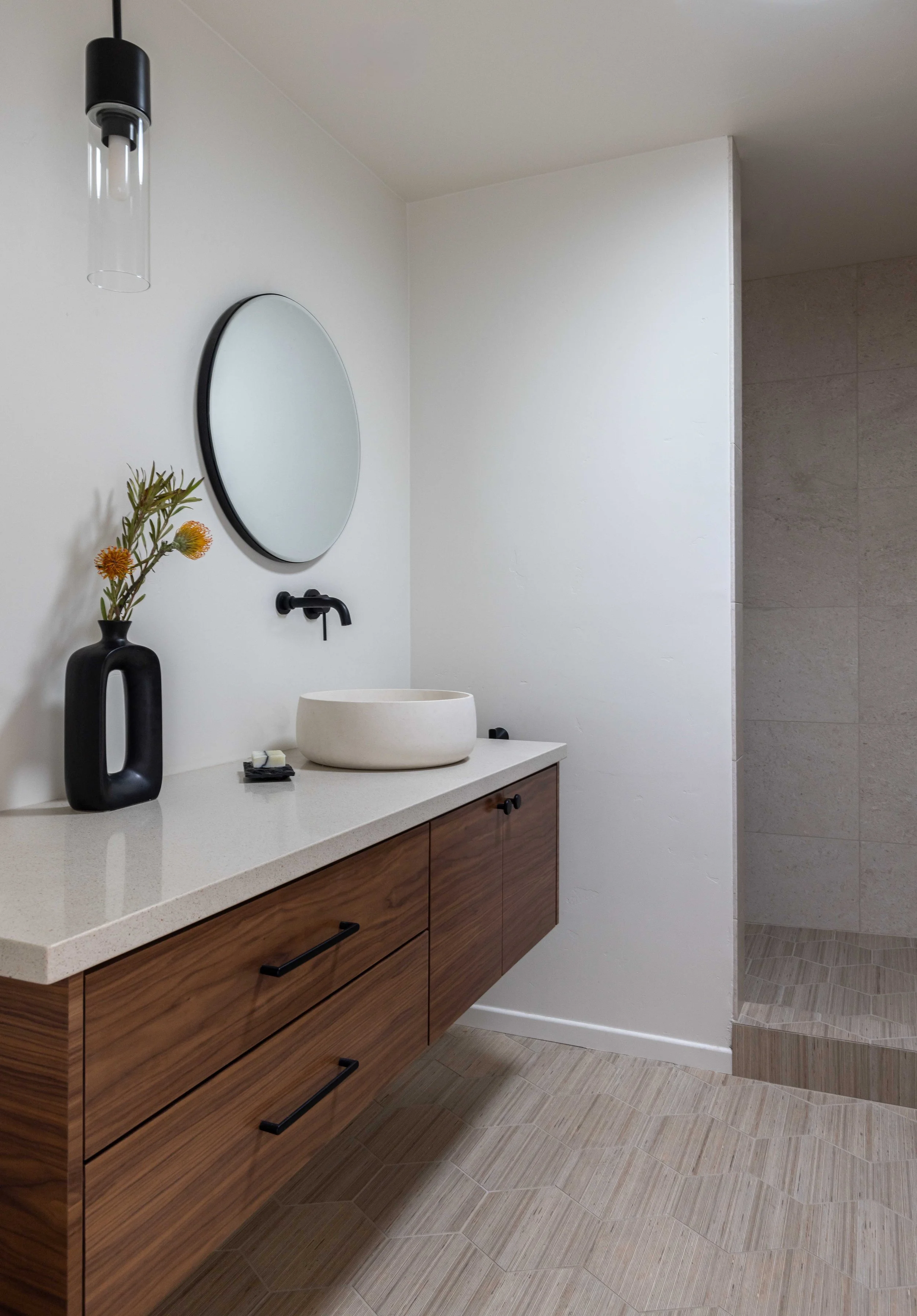 Modern bathroom with a floating wooden vanity, round mirror, vessel sink, and black faucet. Features a black vase with flowers, overhead pendant light, and open shower area.