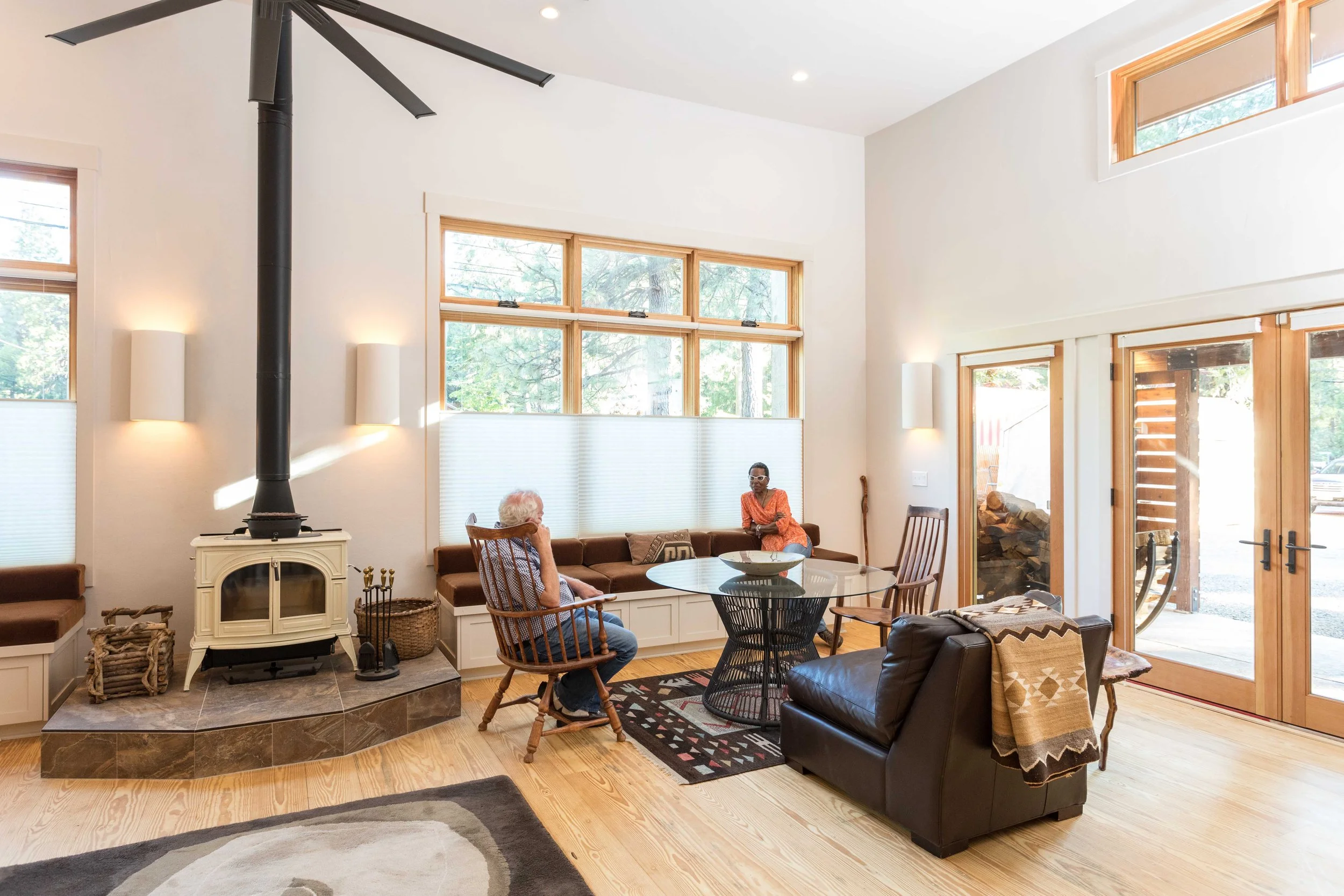 A living room with large windows, a wood-burning stove, two people sitting, wooden chairs, a glass table, and a cozy ambiance. The room has modern decor and natural light.