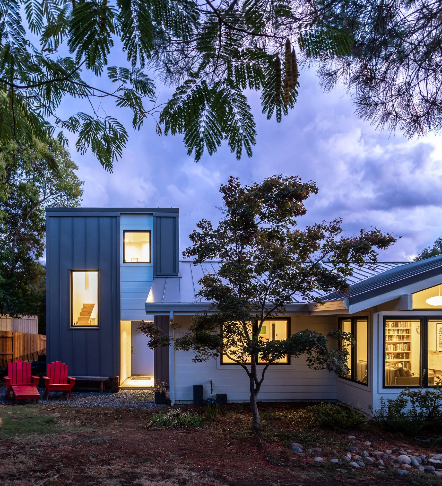 Modern two-story house with lit windows at dusk, featuring a tree in the foreground and red outdoor chairs on the left, surrounded by natural landscaping.