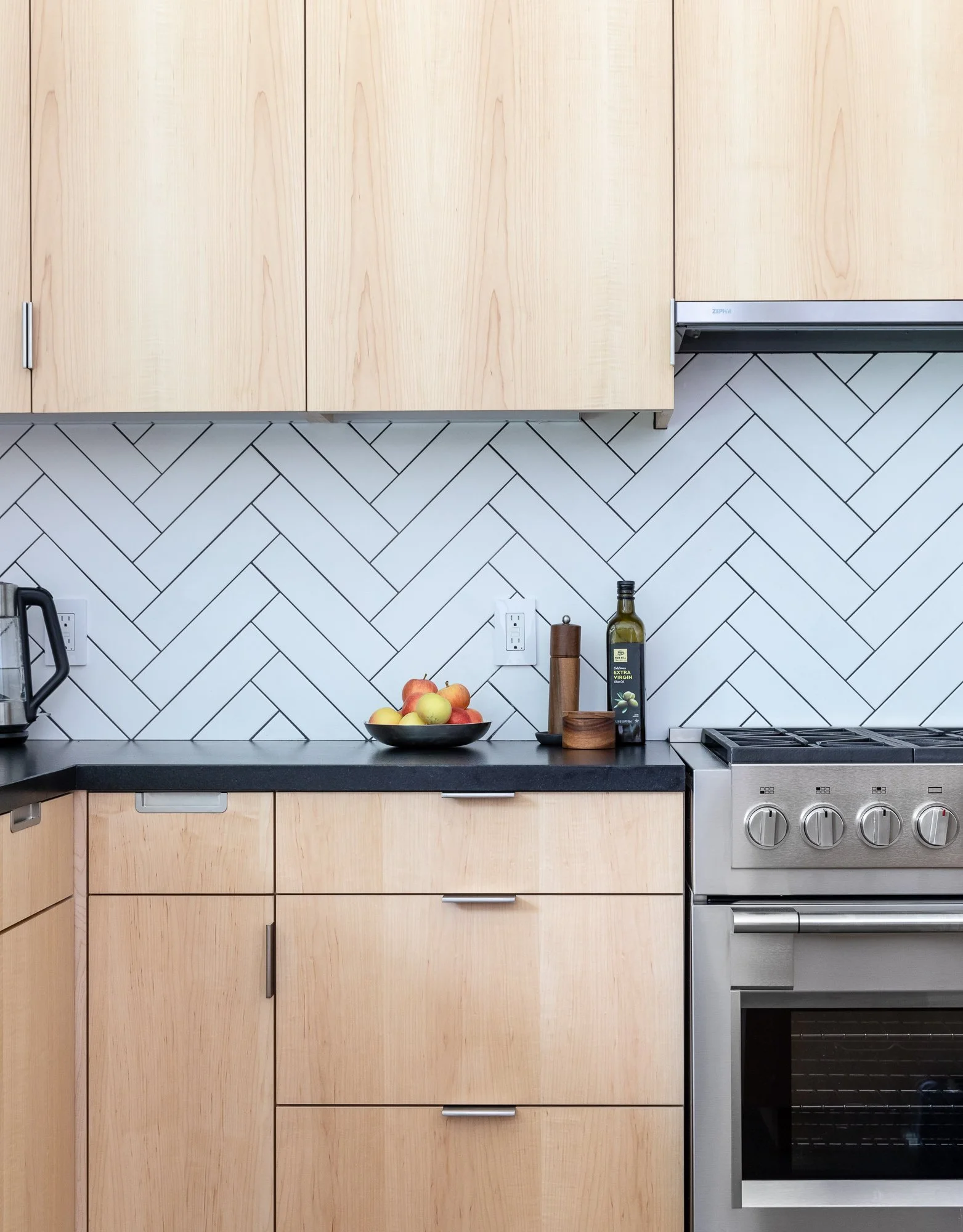 Modern kitchen with light wood cabinets, herringbone tile backsplash, black countertop, and stainless steel stove. A bowl of fruit, olive oil bottle, pepper mill, and kettle are on the counter.