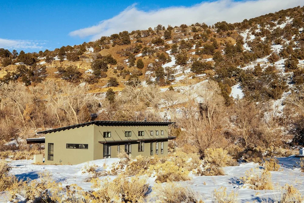 Modern green house with large windows in snowy desert landscape, surrounded by hills and trees.