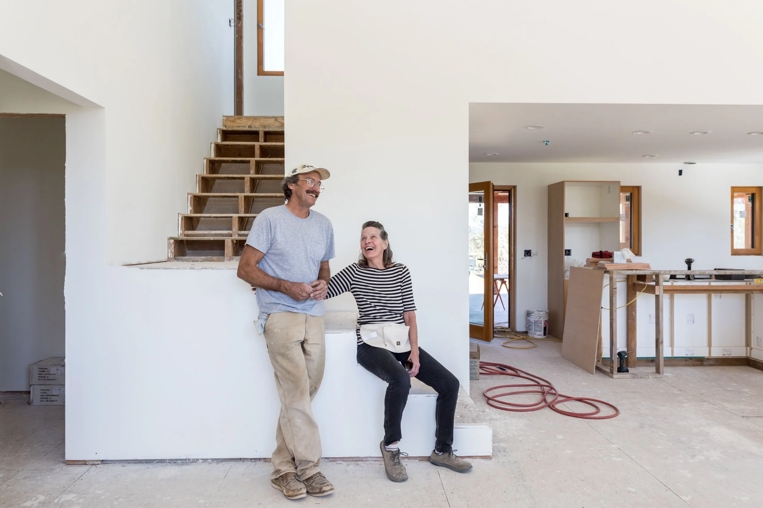 Two people in a partially constructed house, sitting on a ledge near stairs, smiling and talking, with construction materials in the background.