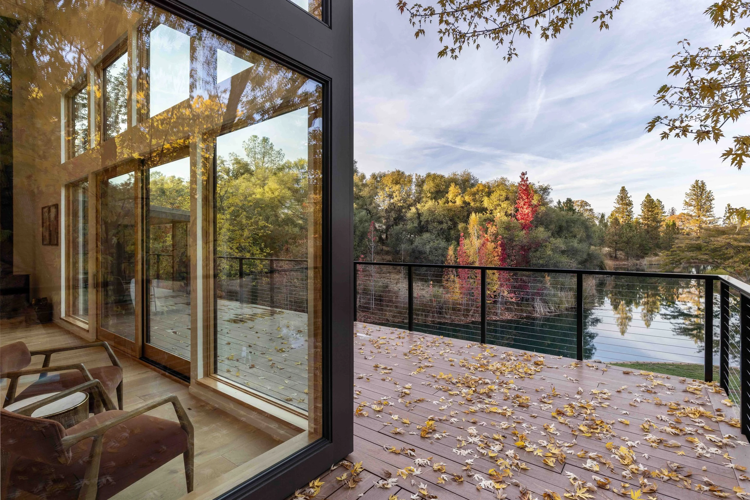 Modern house with large glass windows reflecting trees, with an outdoor deck covered in autumn leaves, overlooking a scenic landscape with trees and a pond.