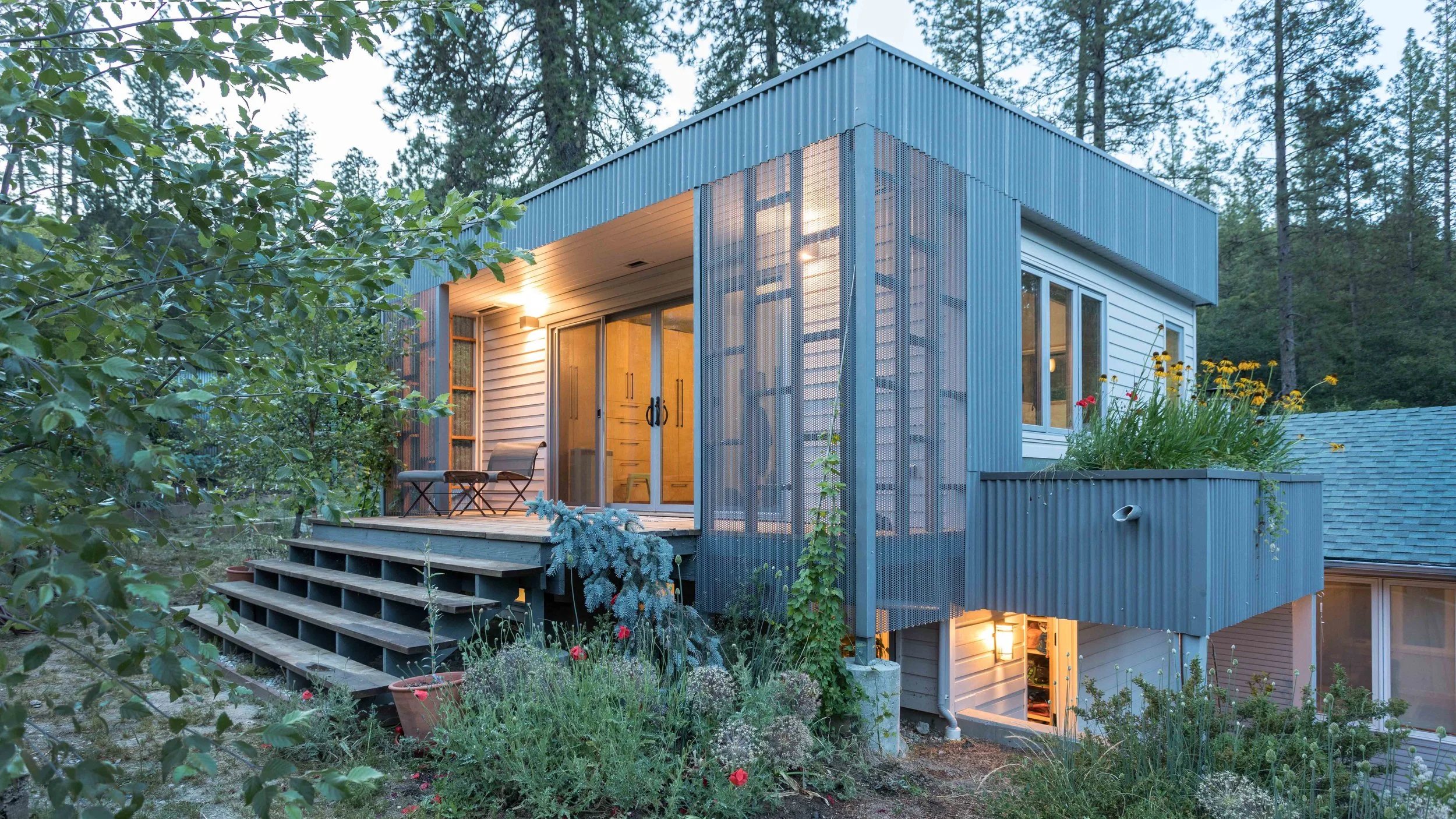 Modern house with metal siding, surrounded by trees, featuring a small porch with steps, outdoor seating, and a vertical garden. Evening lighting creates a warm ambiance.