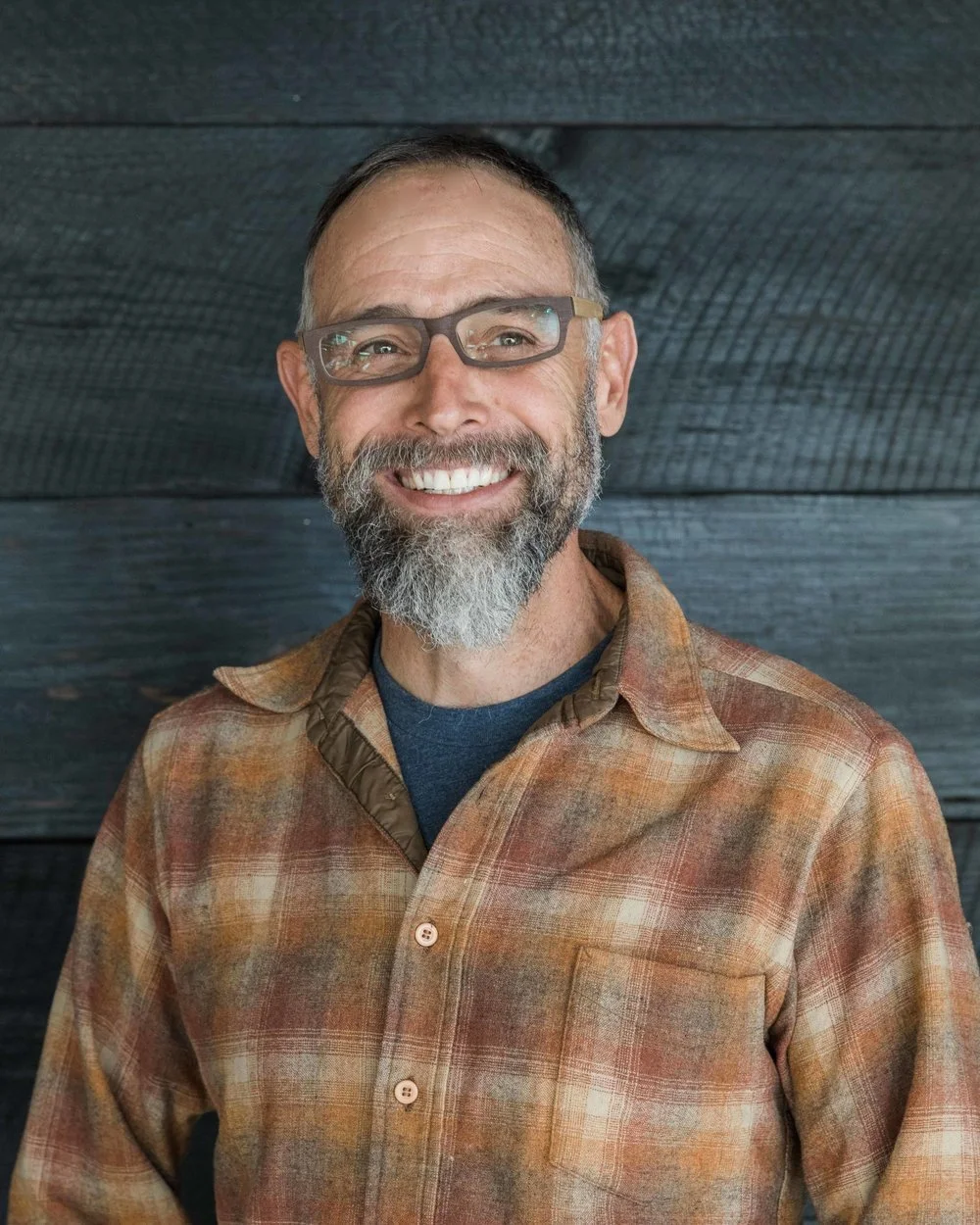 Man with glasses and a beard smiling, wearing a plaid shirt against a dark wood background.